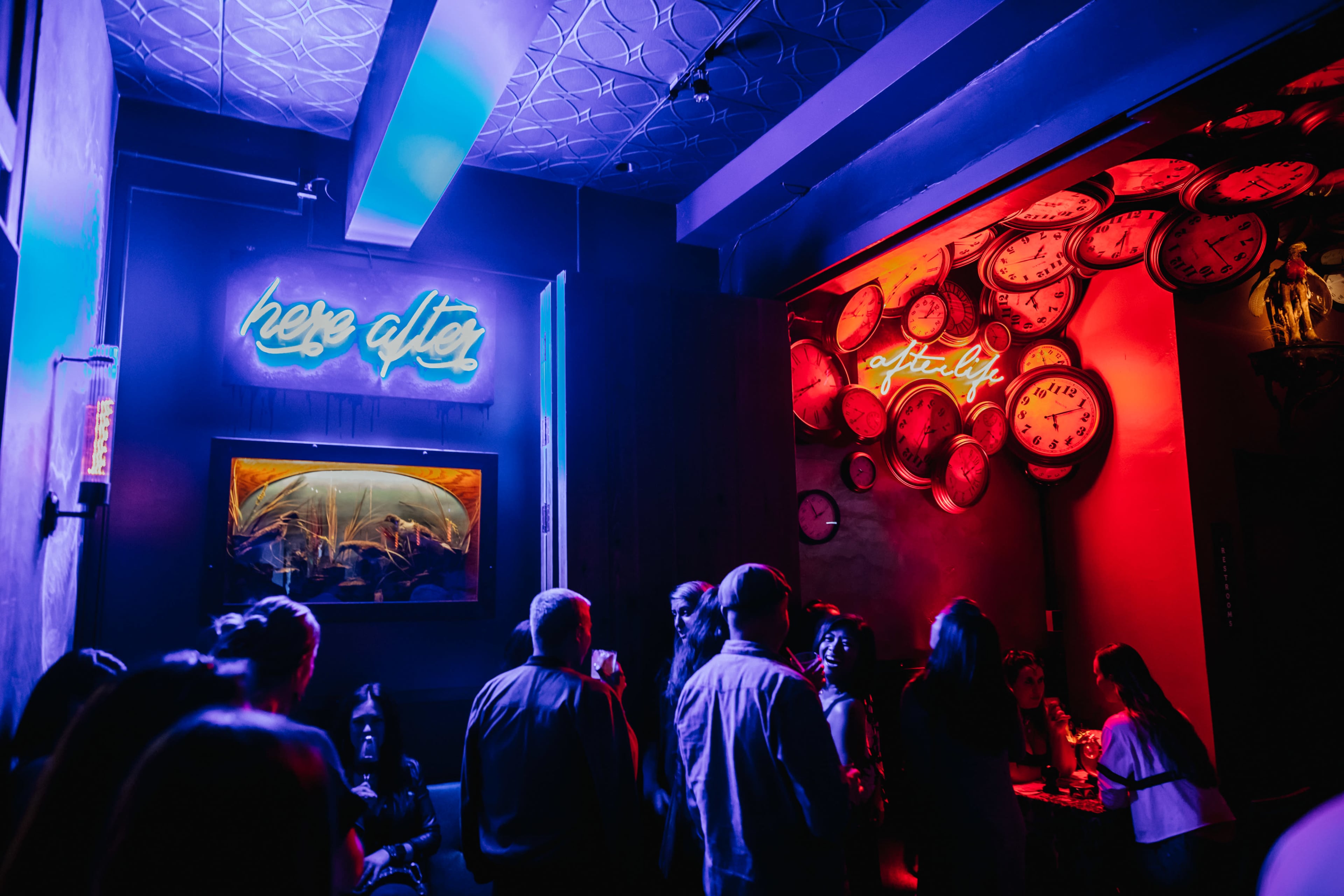 The image shows a dimly lit bar scene with people gathered under neon signs on the walls, one reading "here after" and the other "after life," surrounded by decorative clocks.