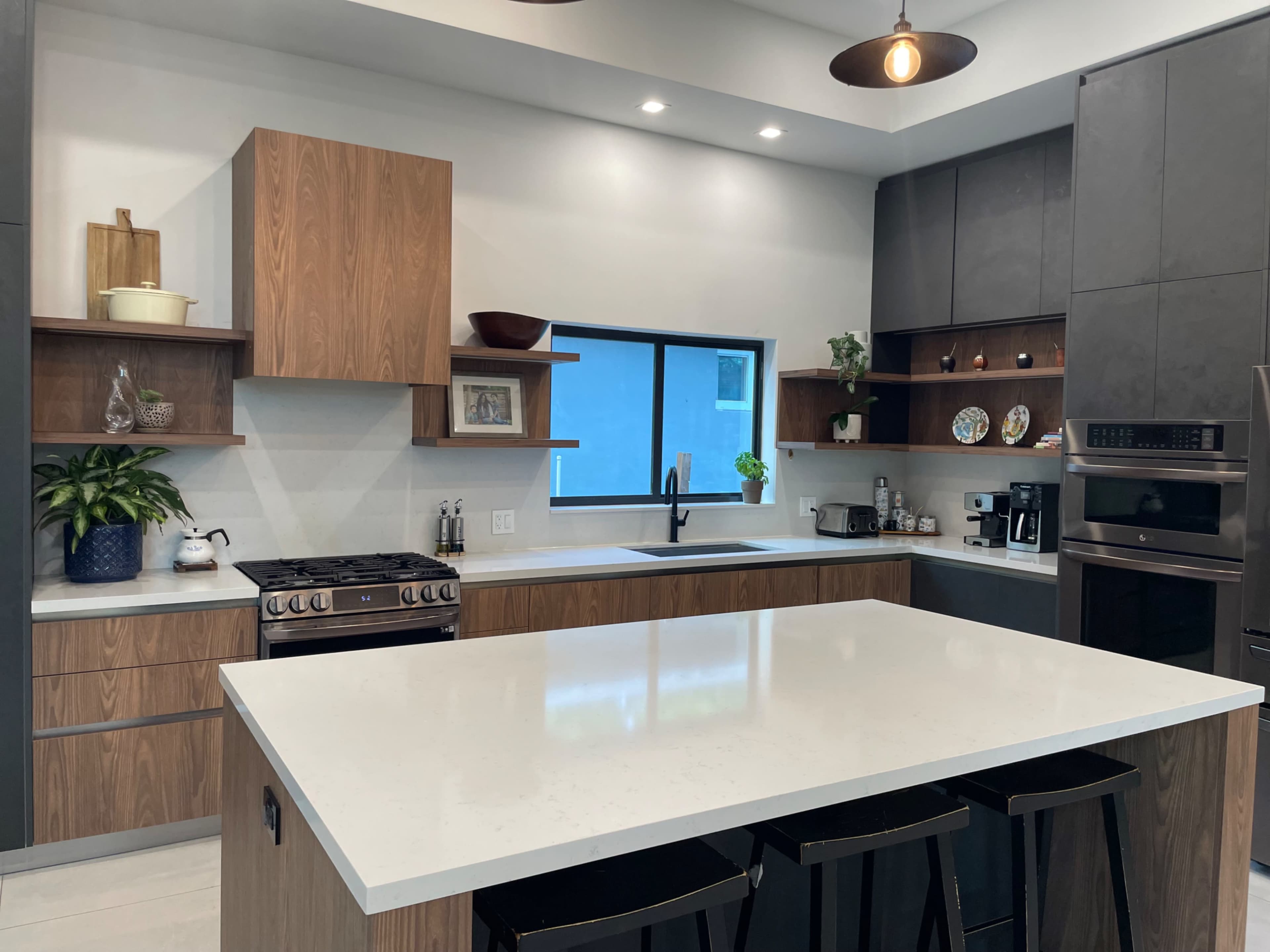 The image shows a modern kitchen with a large central island, stainless steel appliances, wooden cabinetry, and a window providing natural light.