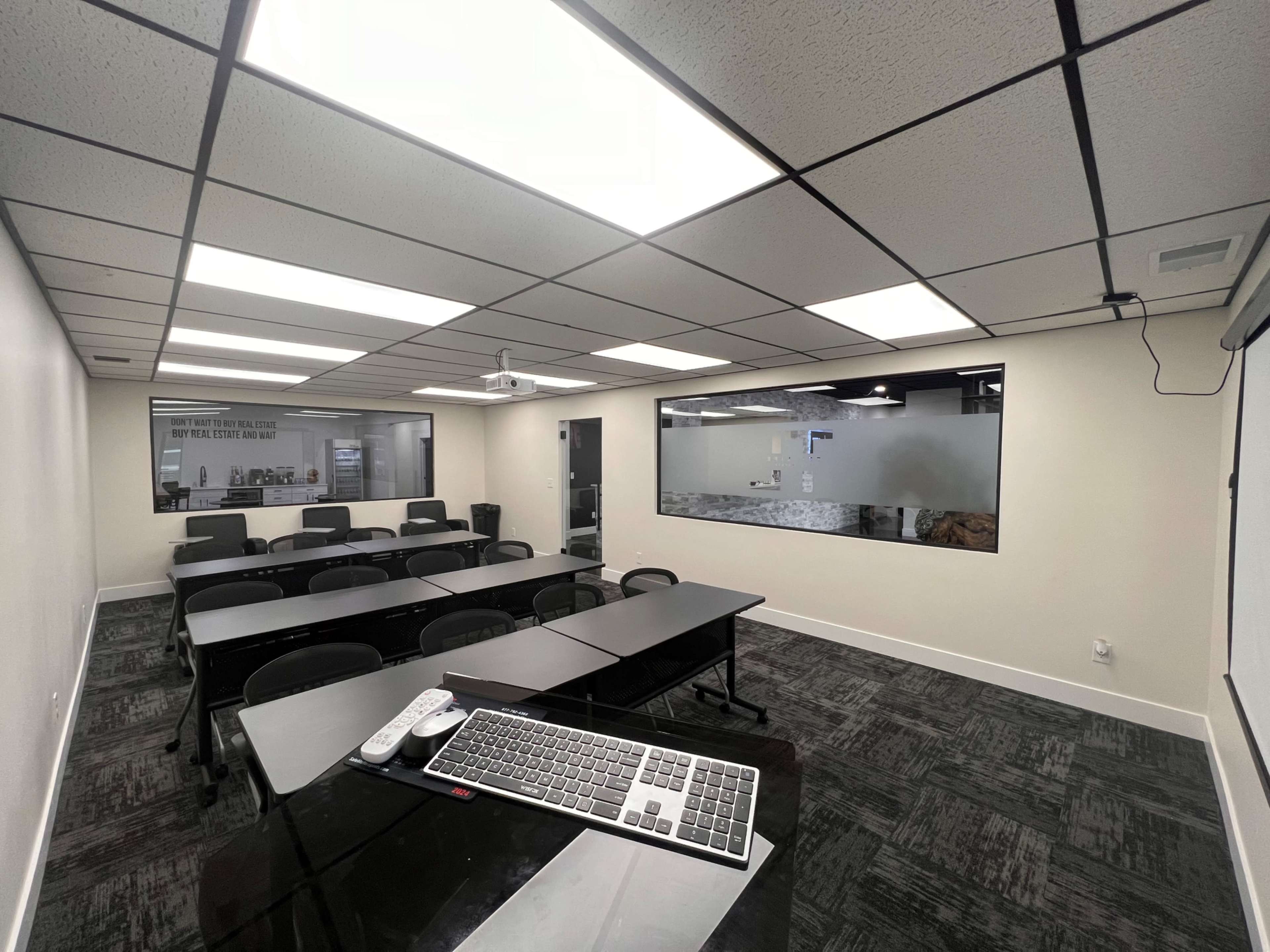 A modern classroom with rows of black chairs and tables, a large screen, and a window displaying a collaborative workspace.