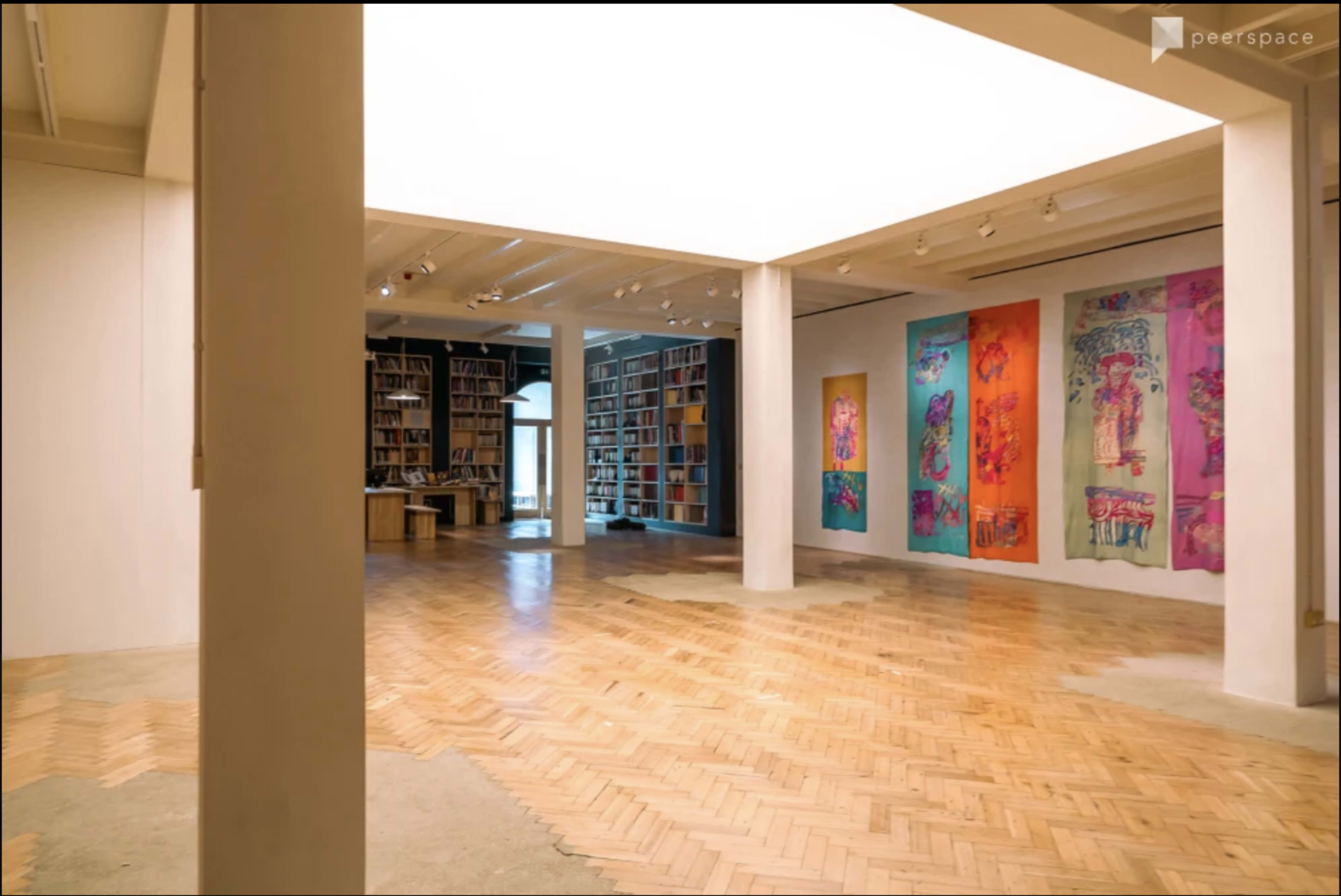 A spacious interior of an art gallery featuring wooden floors, columns, and colorful artworks displayed on the walls.