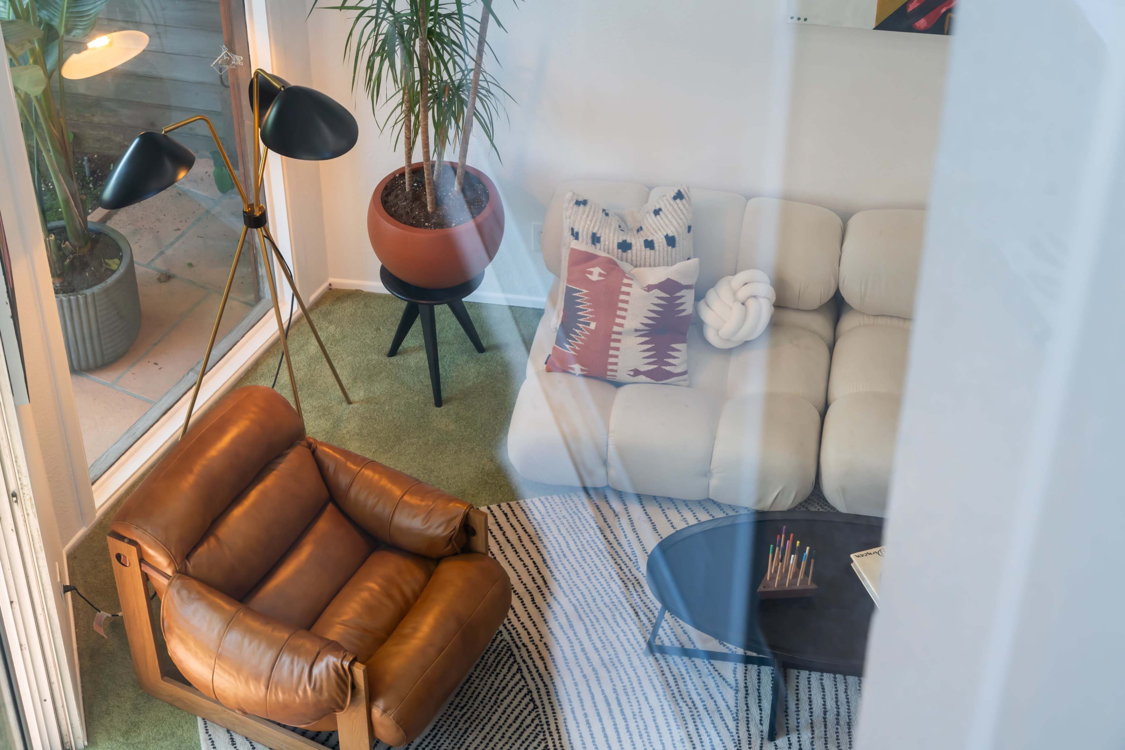 A modern living room with a brown leather chair, a light-colored sofa, a round coffee table with stationery, and potted plants in the background.