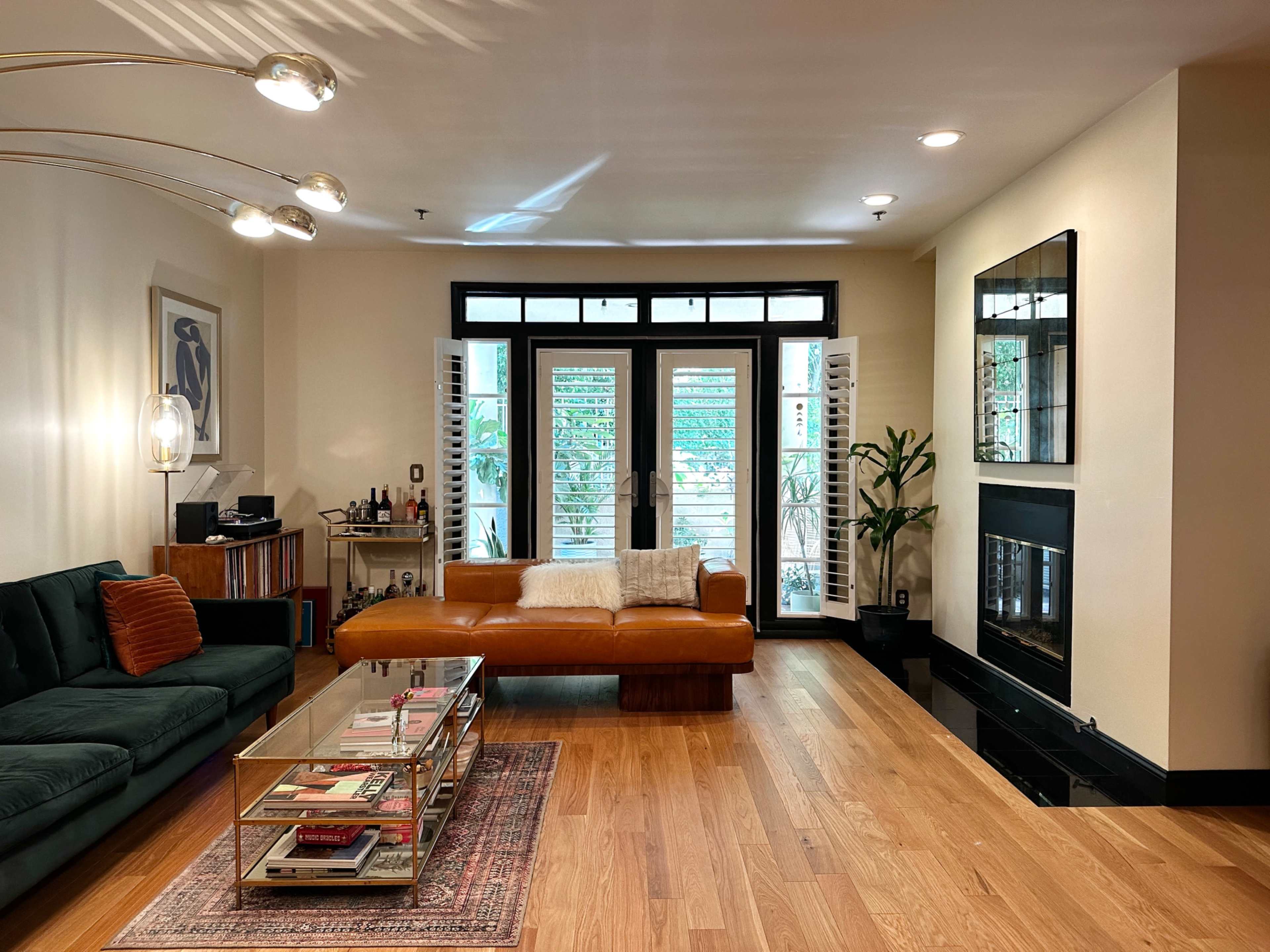 The living room features a green sofa, a brown leather chaise lounge, a glass coffee table with magazines, and a fireplace, with large windows allowing natural light to enter.