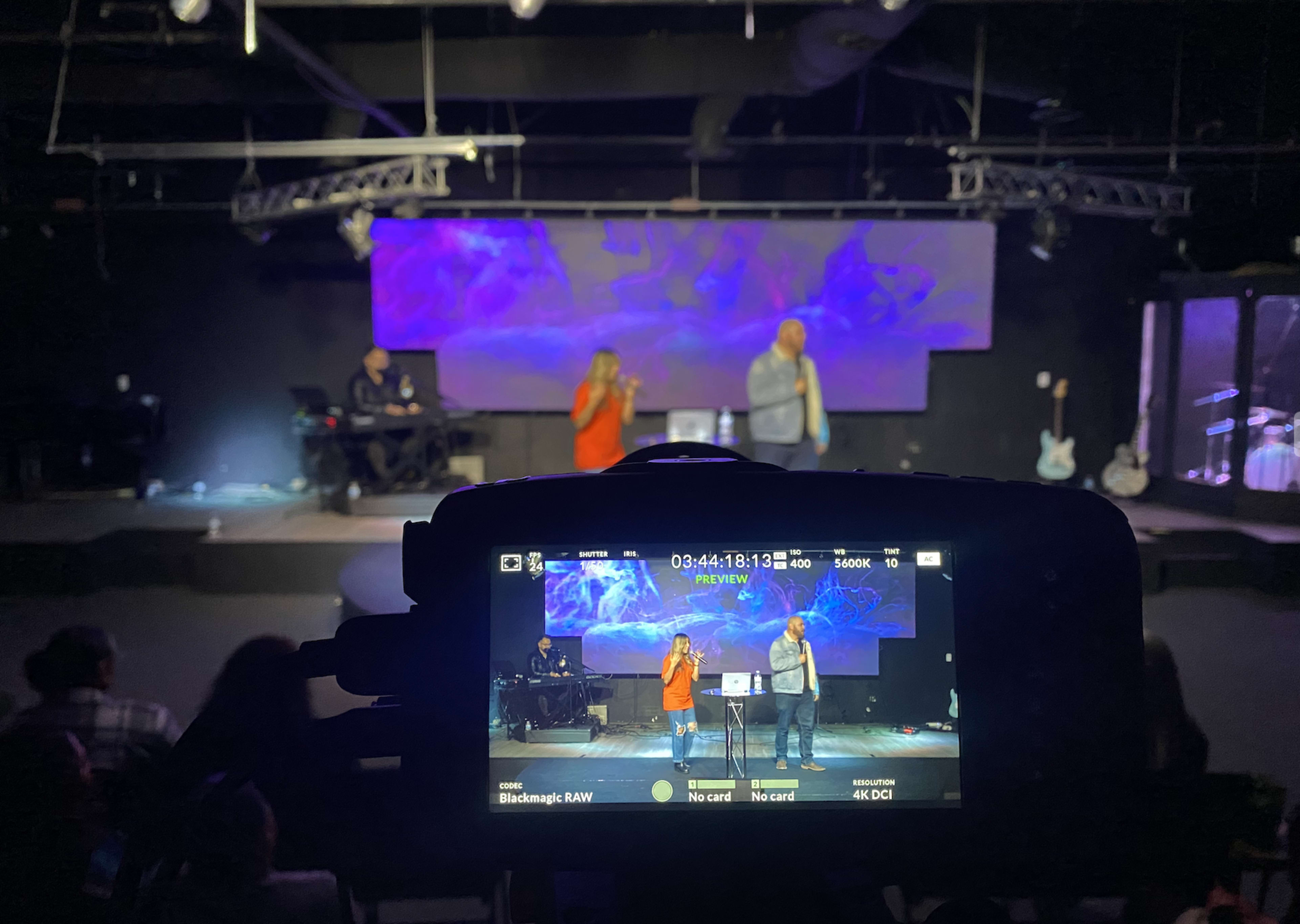 A person is speaking on stage in front of an audience while another person stands beside them, with a live band in the background and a video camera capturing the scene.