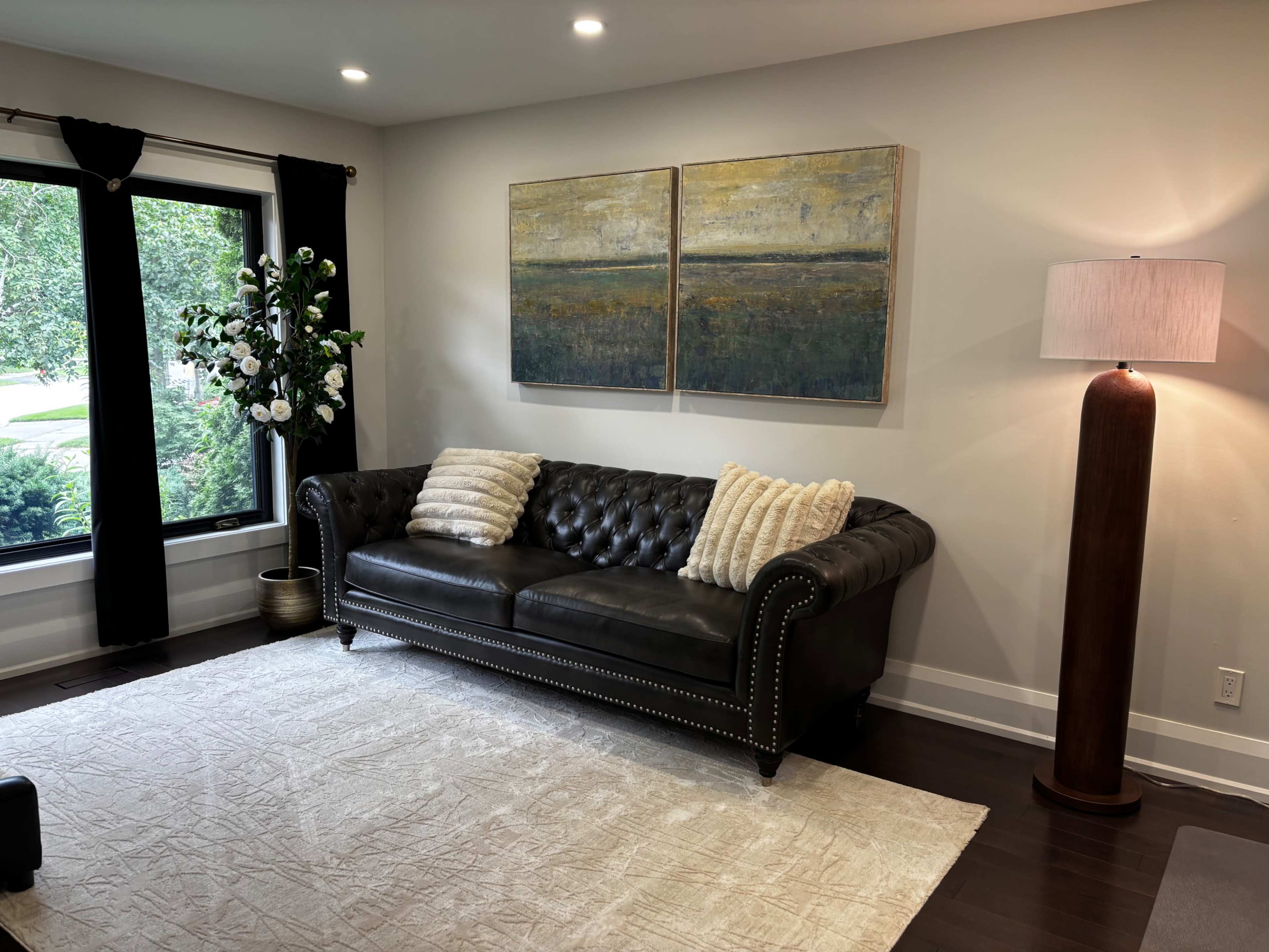 The image shows a living room with a dark leather sofa adorned with decorative pillows, a large abstract painting on the wall, and a standing lamp next to a window with curtains.