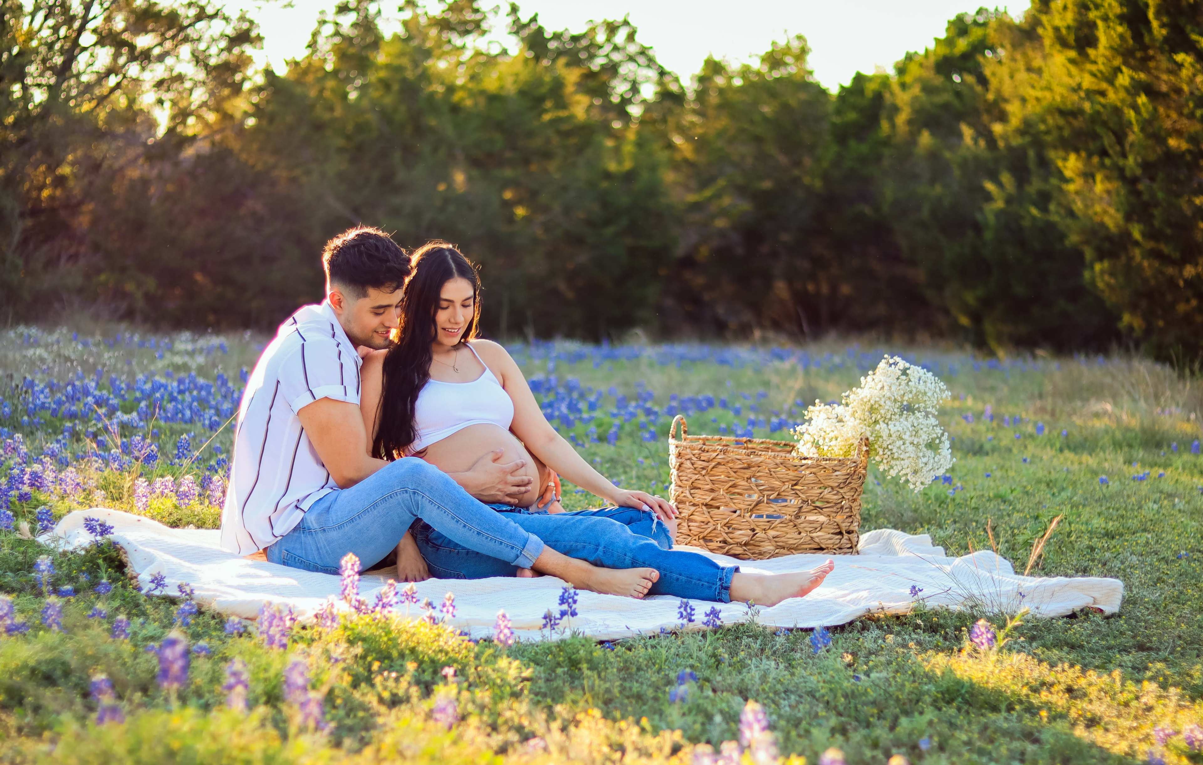 A couple sits on a blanket in a field of bluebonnets, with the woman cradling her pregnant belly and a basket of flowers beside them.