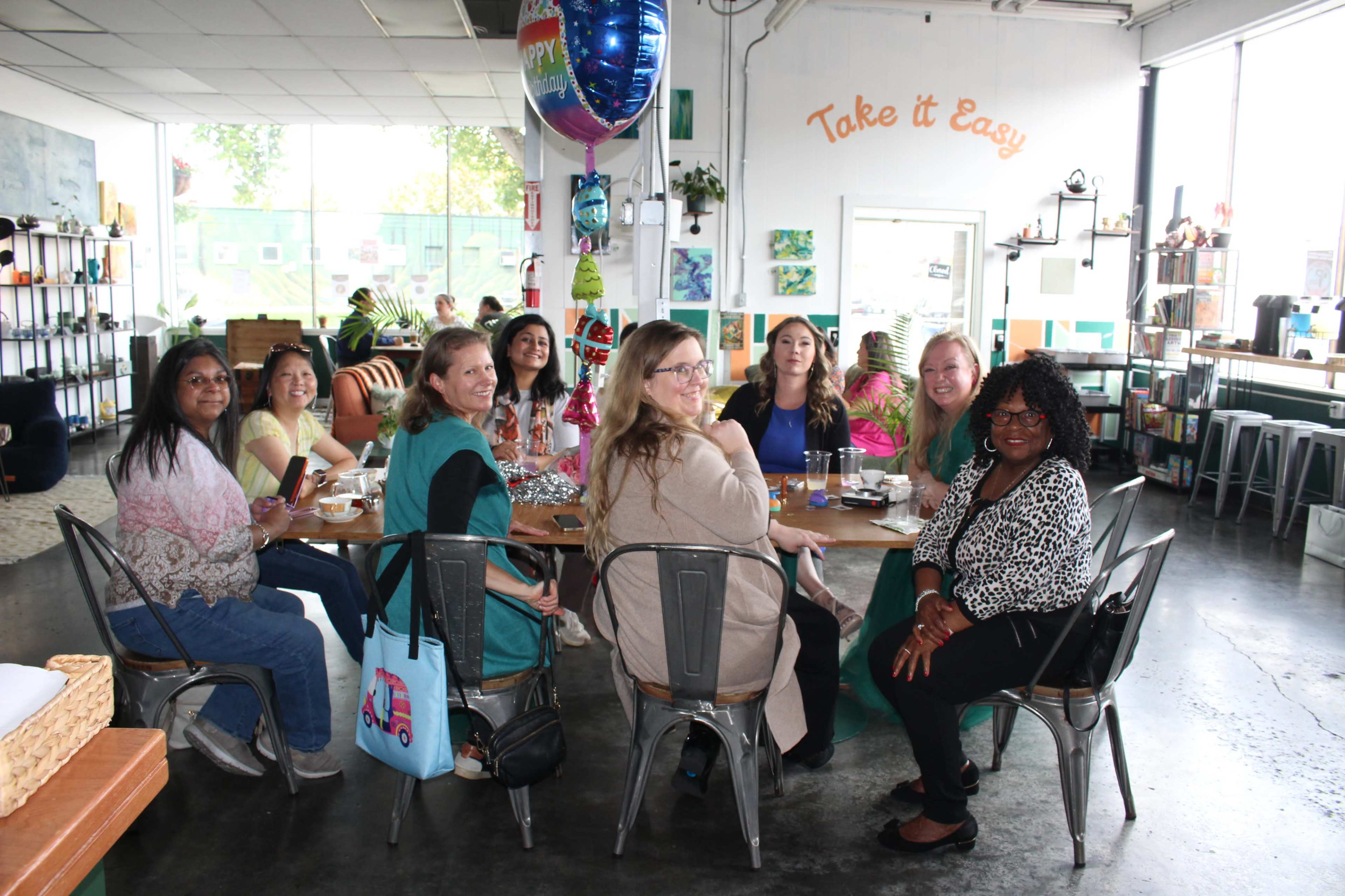 A group of ten women is seated at a long table in a brightly lit cafe, with decorations and balloons in the background.