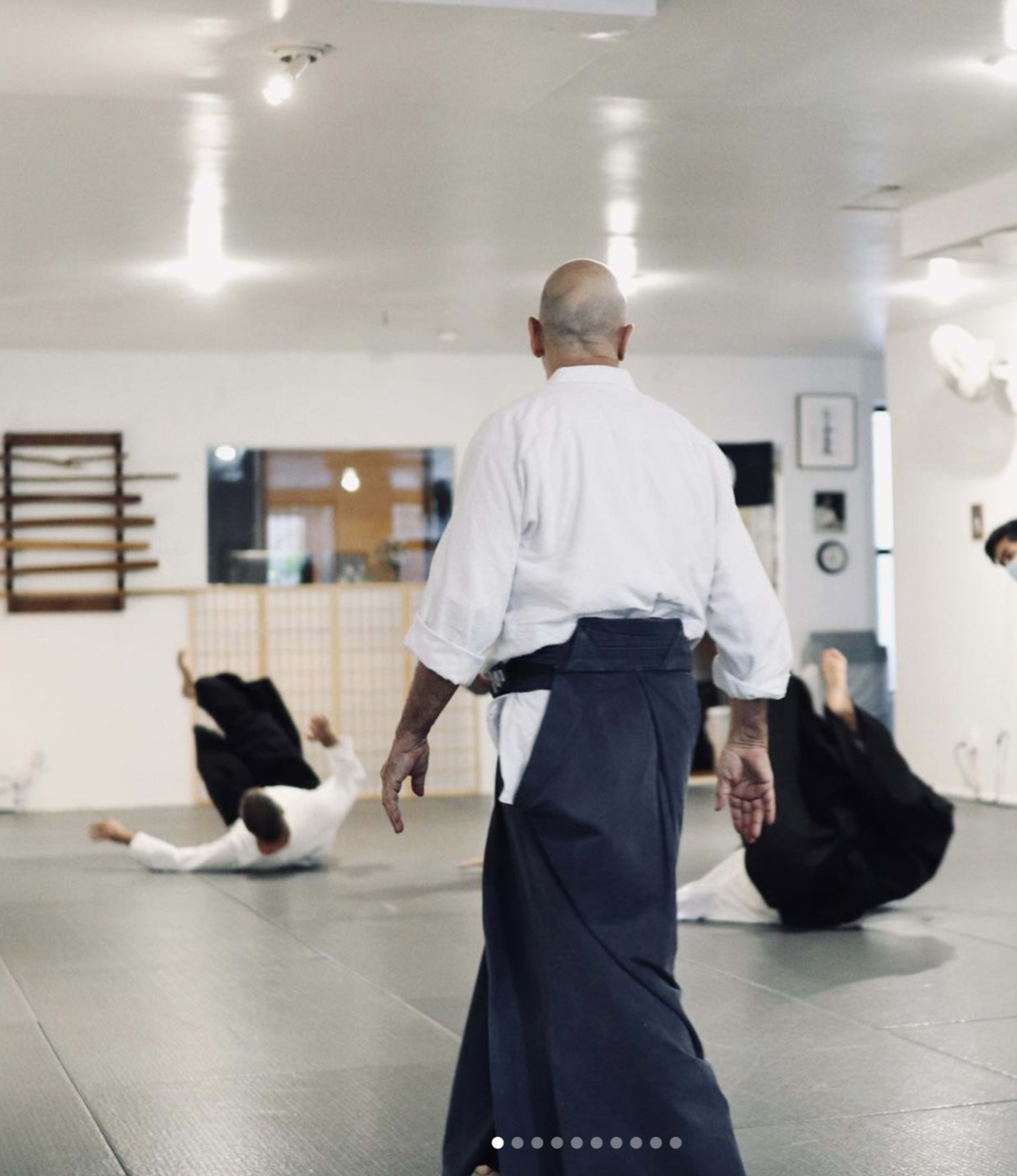 A martial arts instructor observes students practicing on a mat in a training studio.