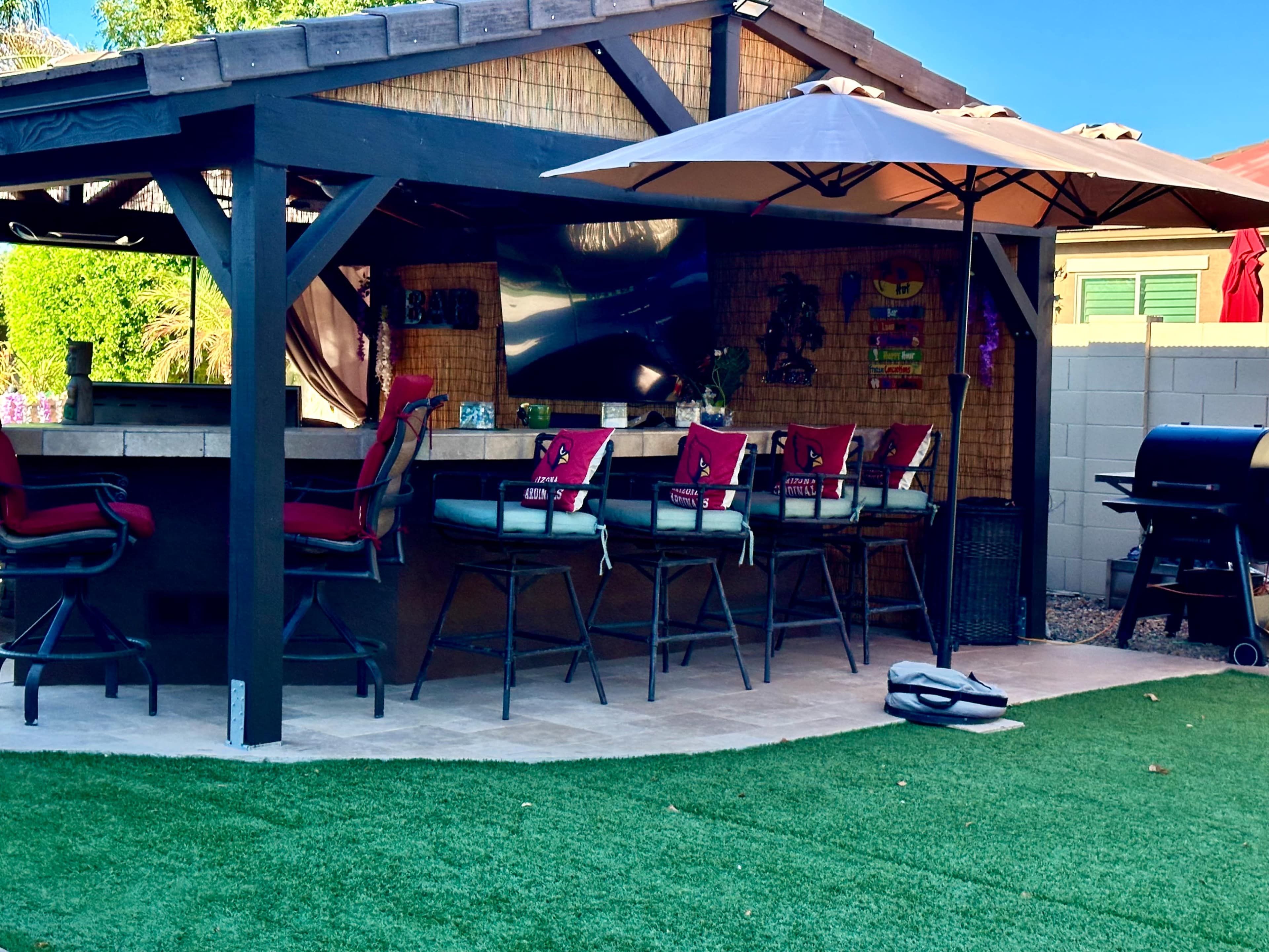 A backyard bar area with high stools, an umbrella, and a grill, surrounded by artificial turf.