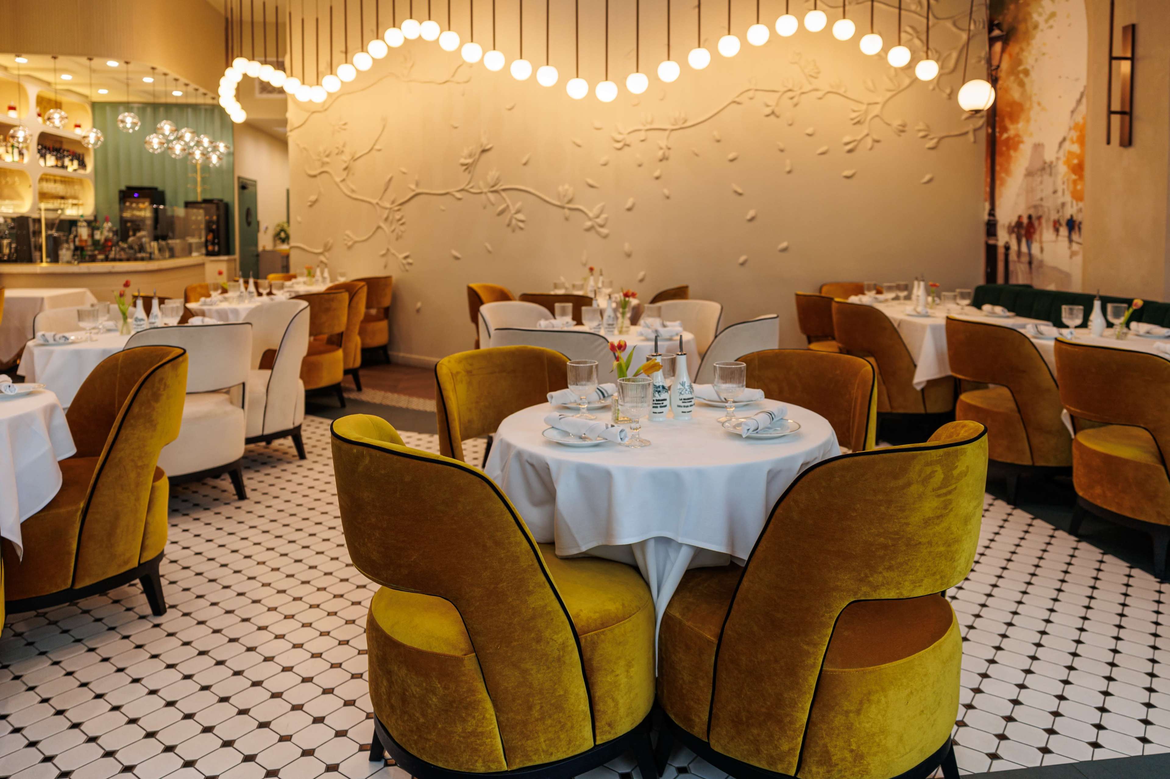 The image shows an elegantly designed restaurant interior with round tables set for dining, featuring yellow velvet chairs and a decorative wall with abstract designs.