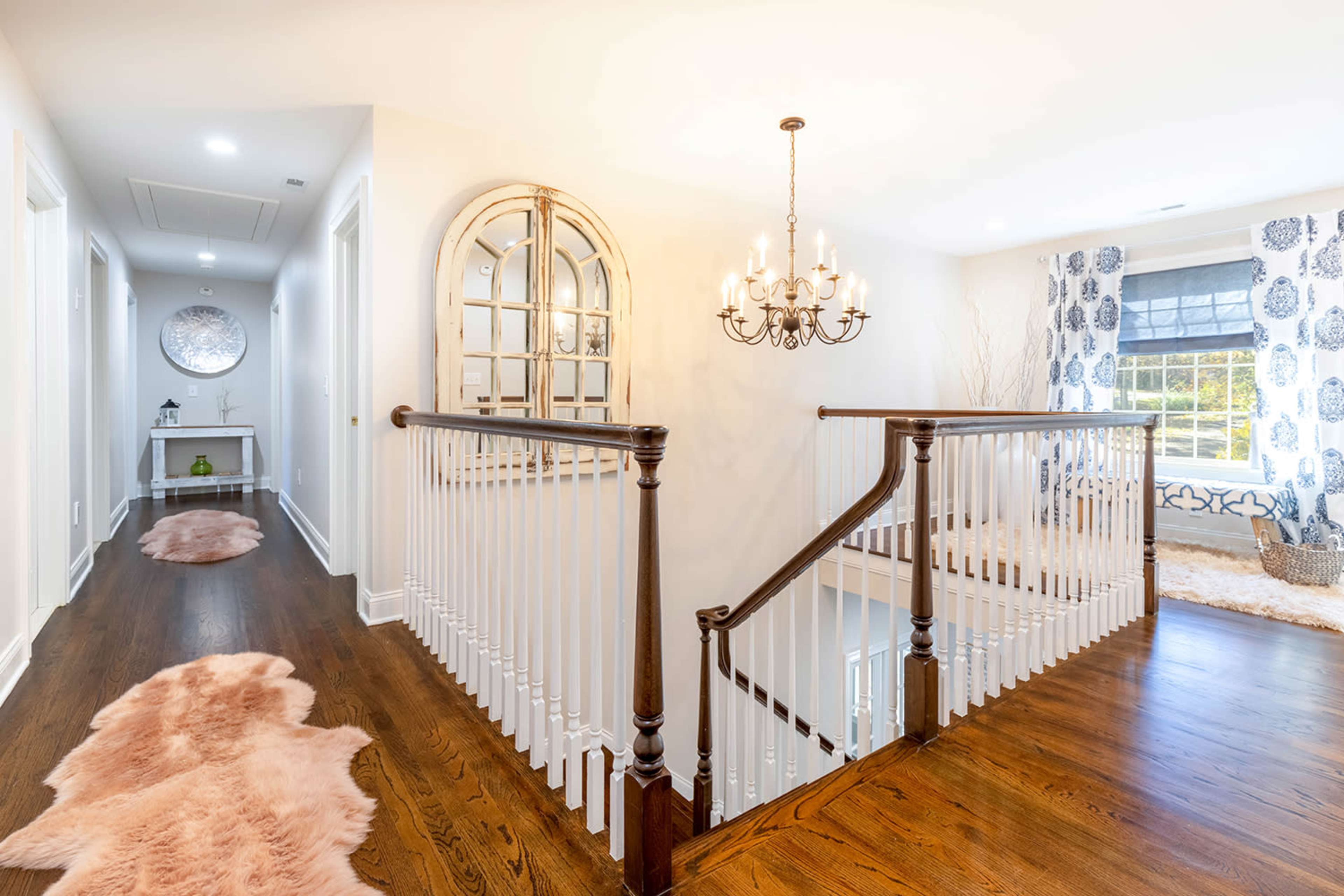 The image shows a well-lit hallway with a wooden staircase, a decorative mirror, and a chandelier above, featuring a plush rug and curtains with a floral pattern in the background.