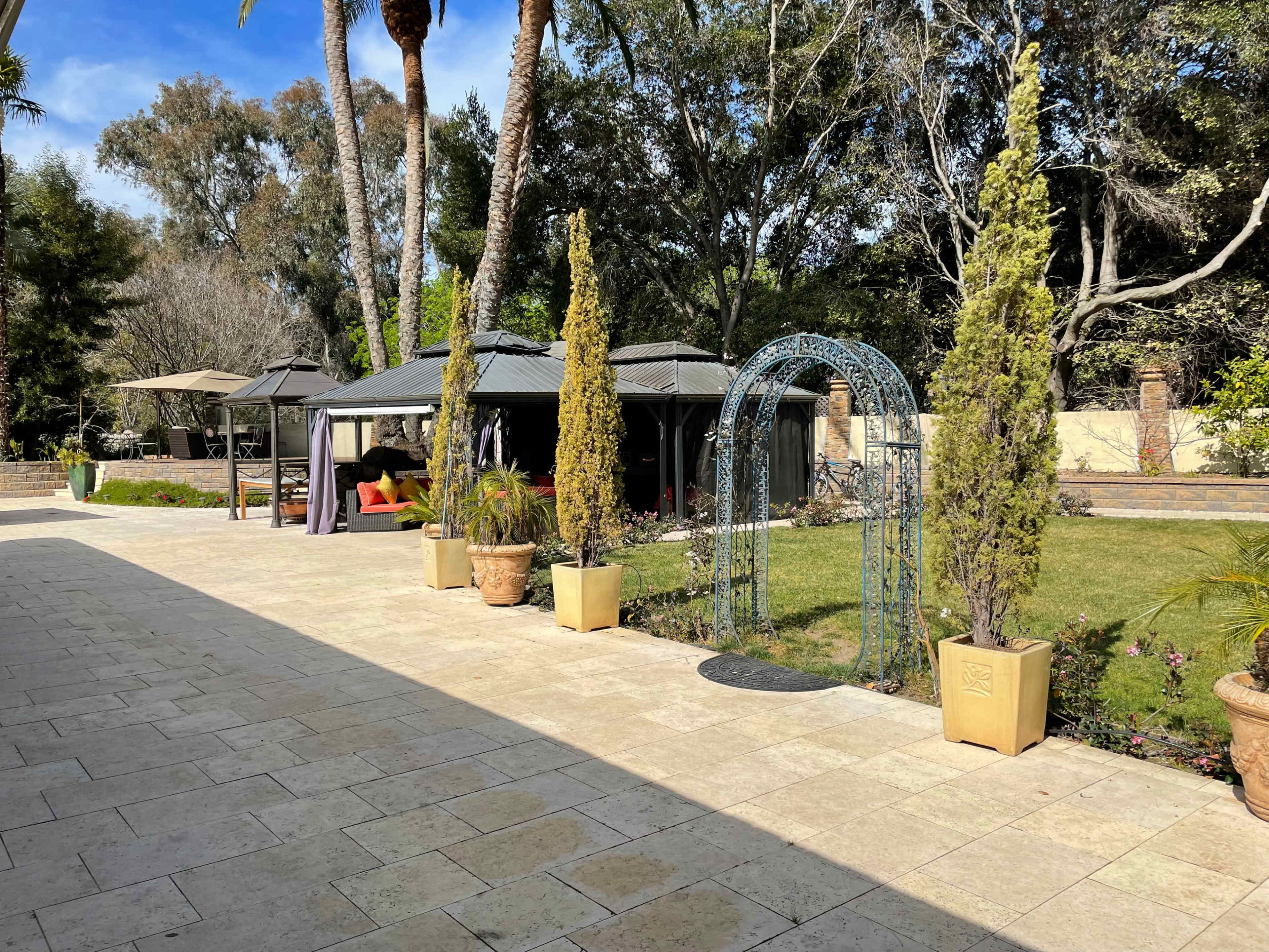 The image shows a landscaped backyard featuring potted plants, a gazebo, and a decorative archway amidst grassy areas and palm trees.