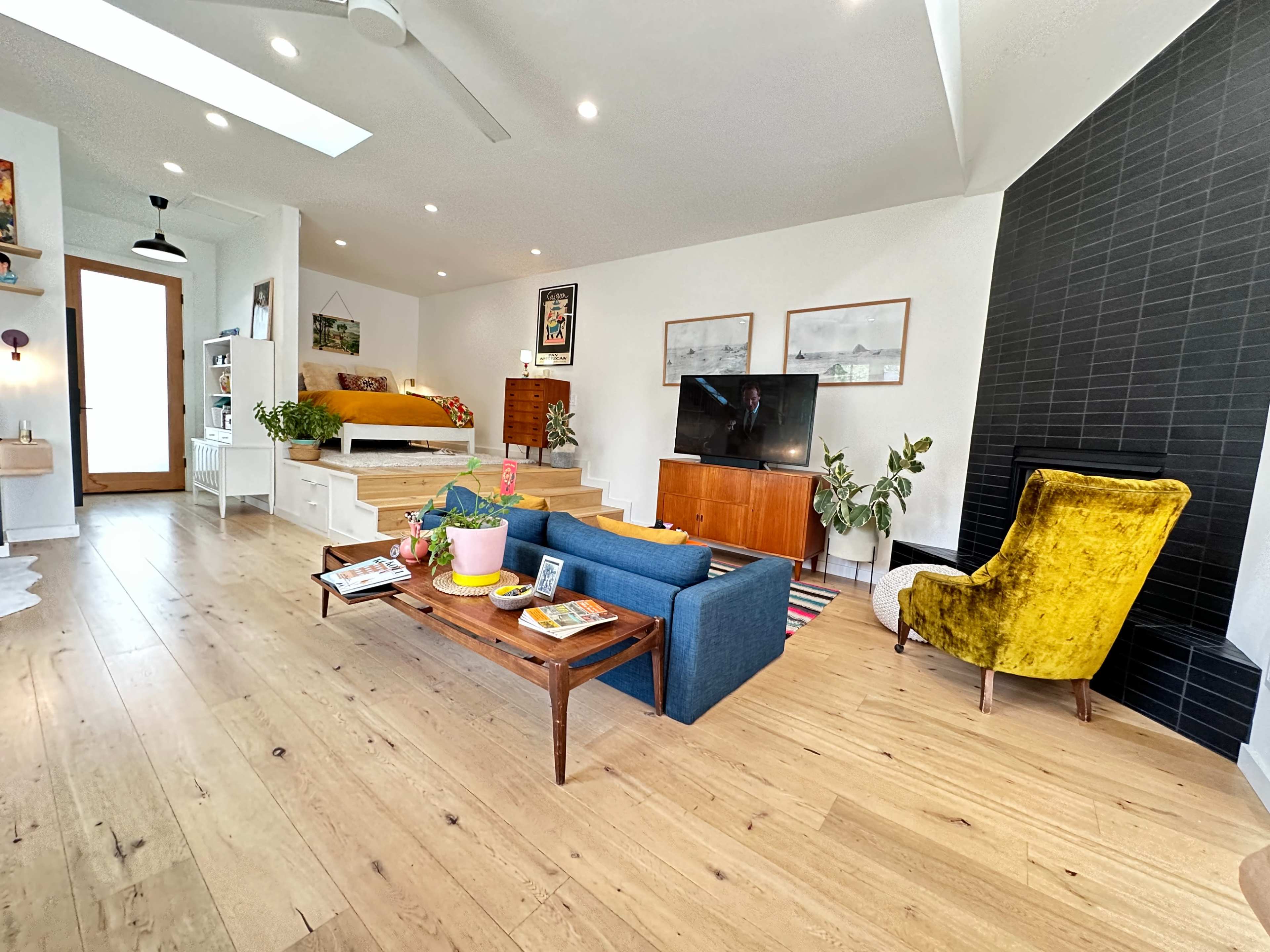 A modern living room with a wooden floor, a blue sofa, a yellow armchair, a coffee table with magazines, and a large television against a black wall.