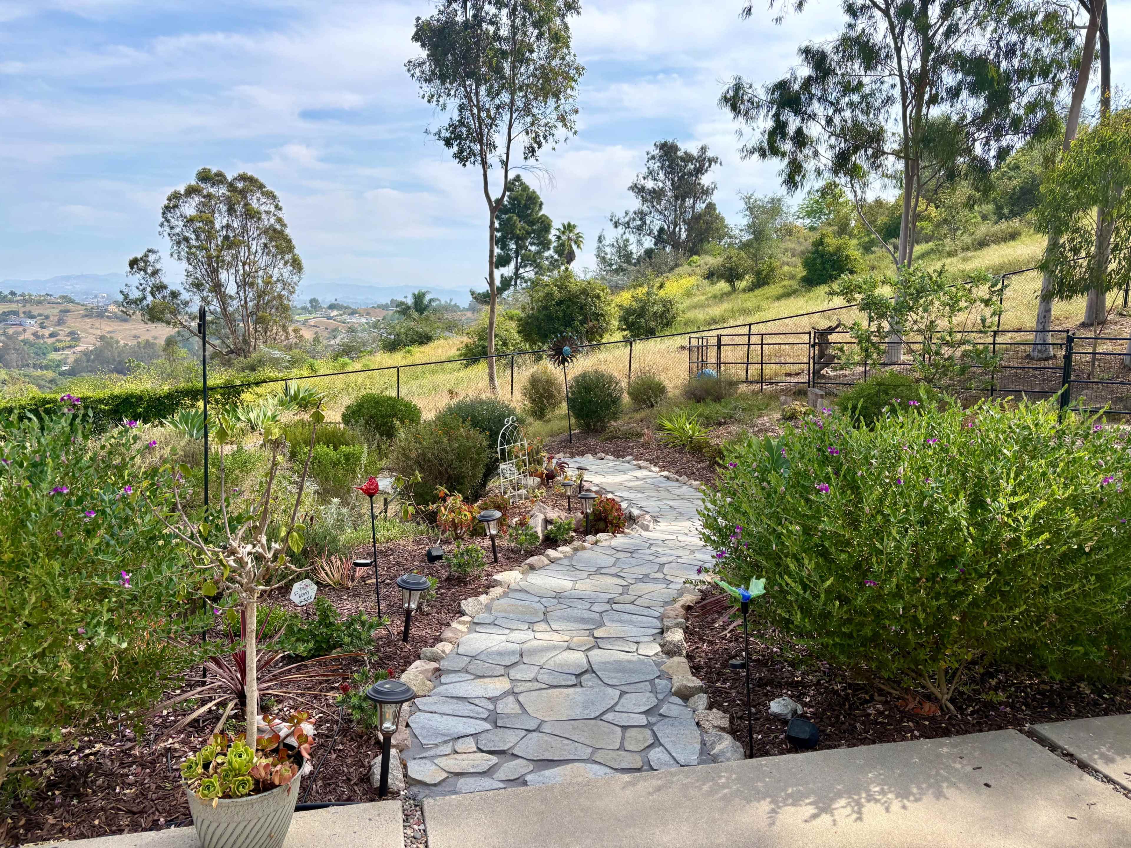 A stone pathway winds through a lush garden, leading to a hillside with distant hills and trees under a partly cloudy sky.