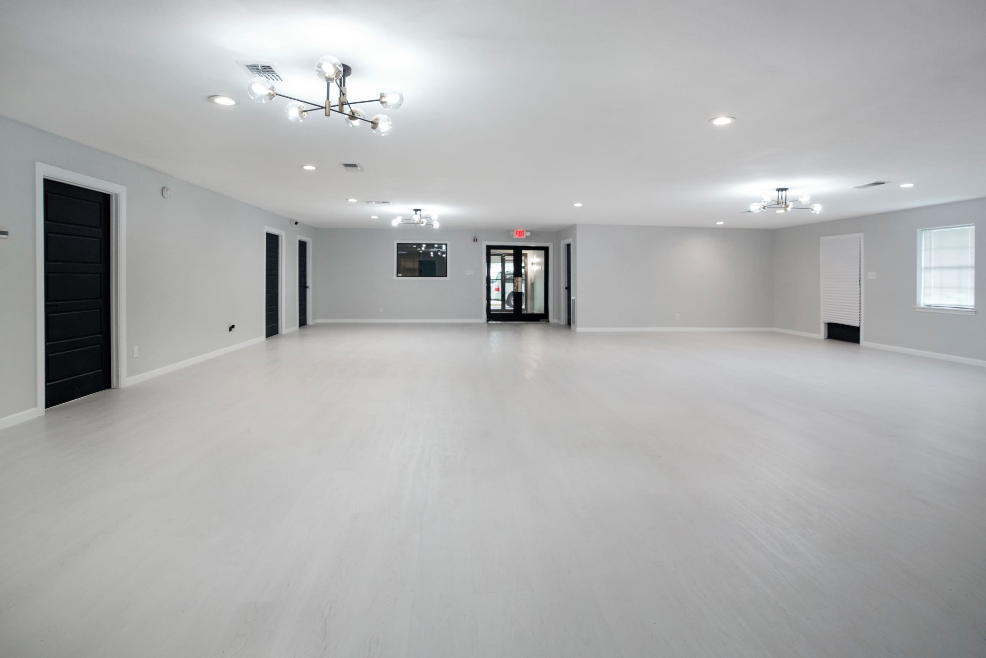 The image displays a spacious, empty room with light gray walls and a white floor, featuring several light fixtures and black doors leading to other areas.