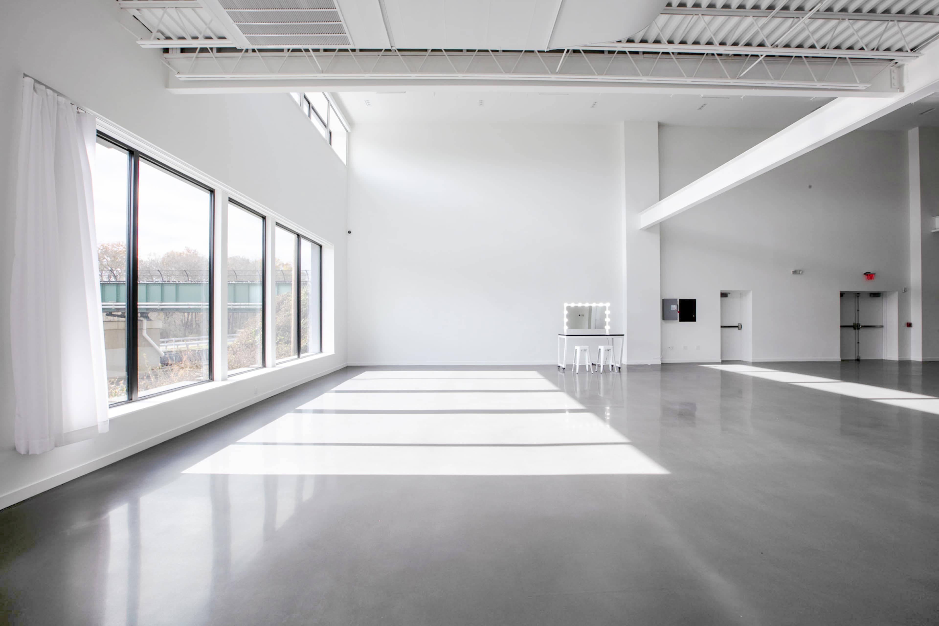 A spacious, well-lit interior with large windows, concrete flooring, and a vanity table near the windows.