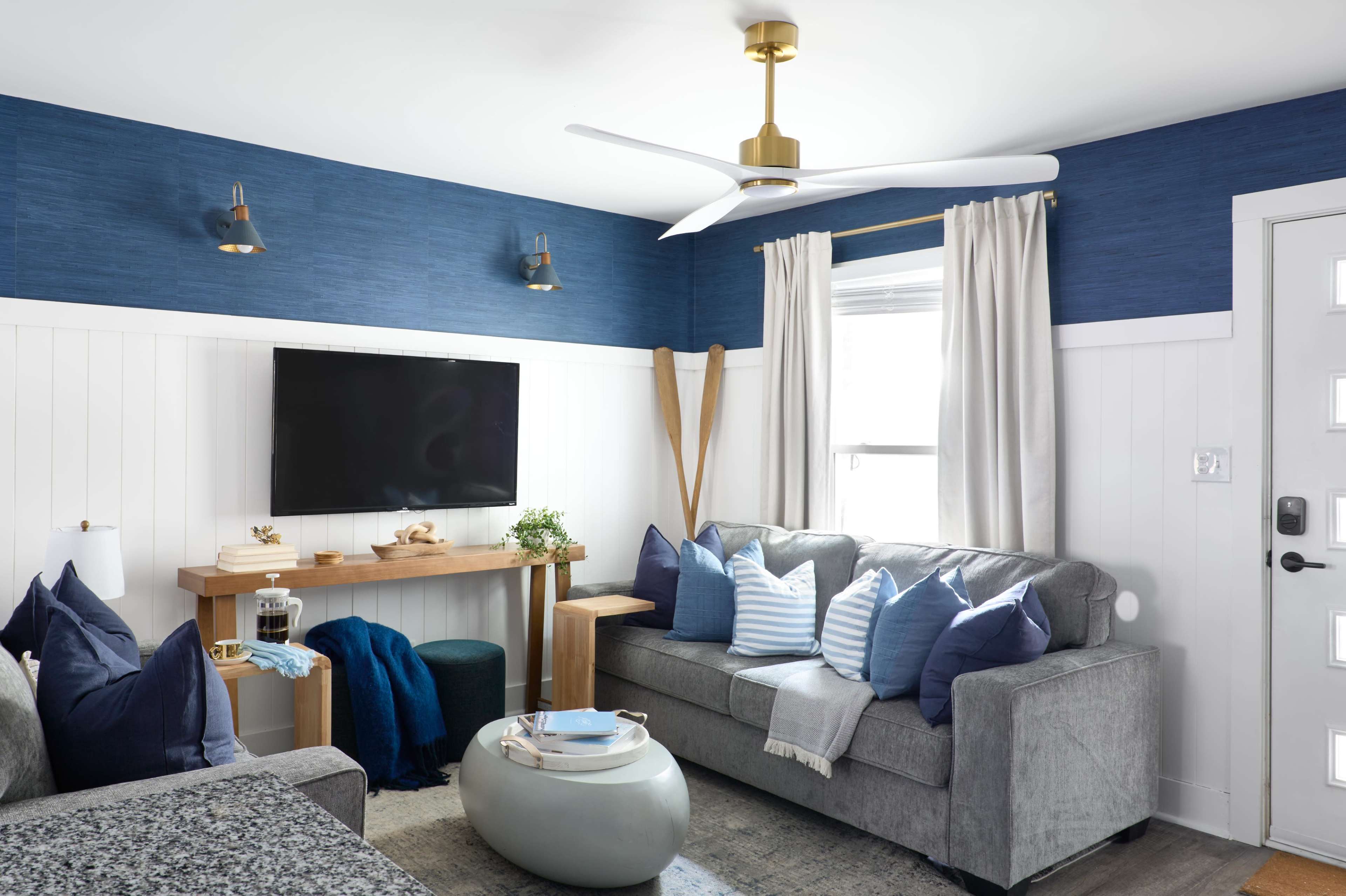 The image shows a modern living room featuring a gray sofa with blue and white pillows, a wall-mounted television, and a wooden shelf displaying decor items, all complemented by blue walls and natural light from a window.