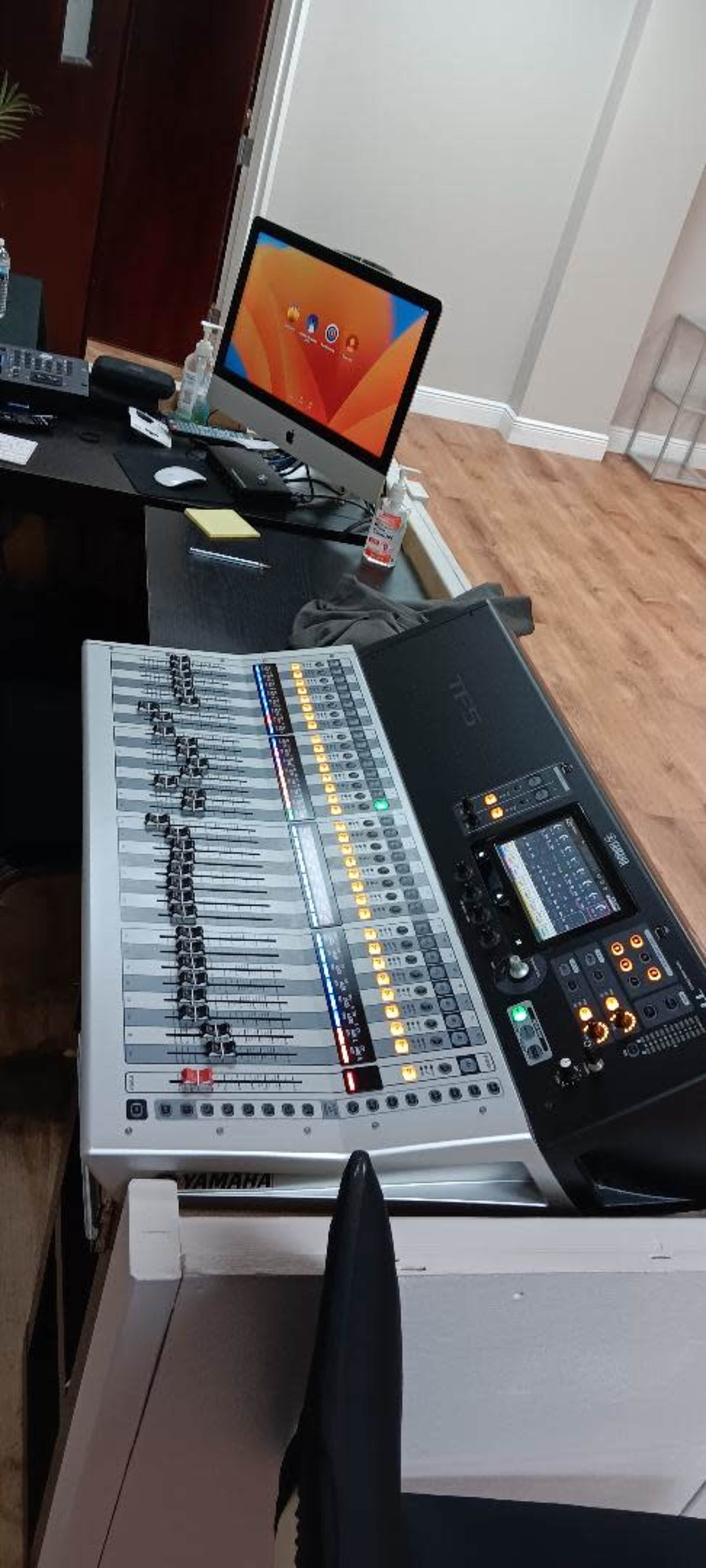 The image shows a Yamaha digital audio mixer on a table next to an iMac computer in an office setting with wooden flooring.