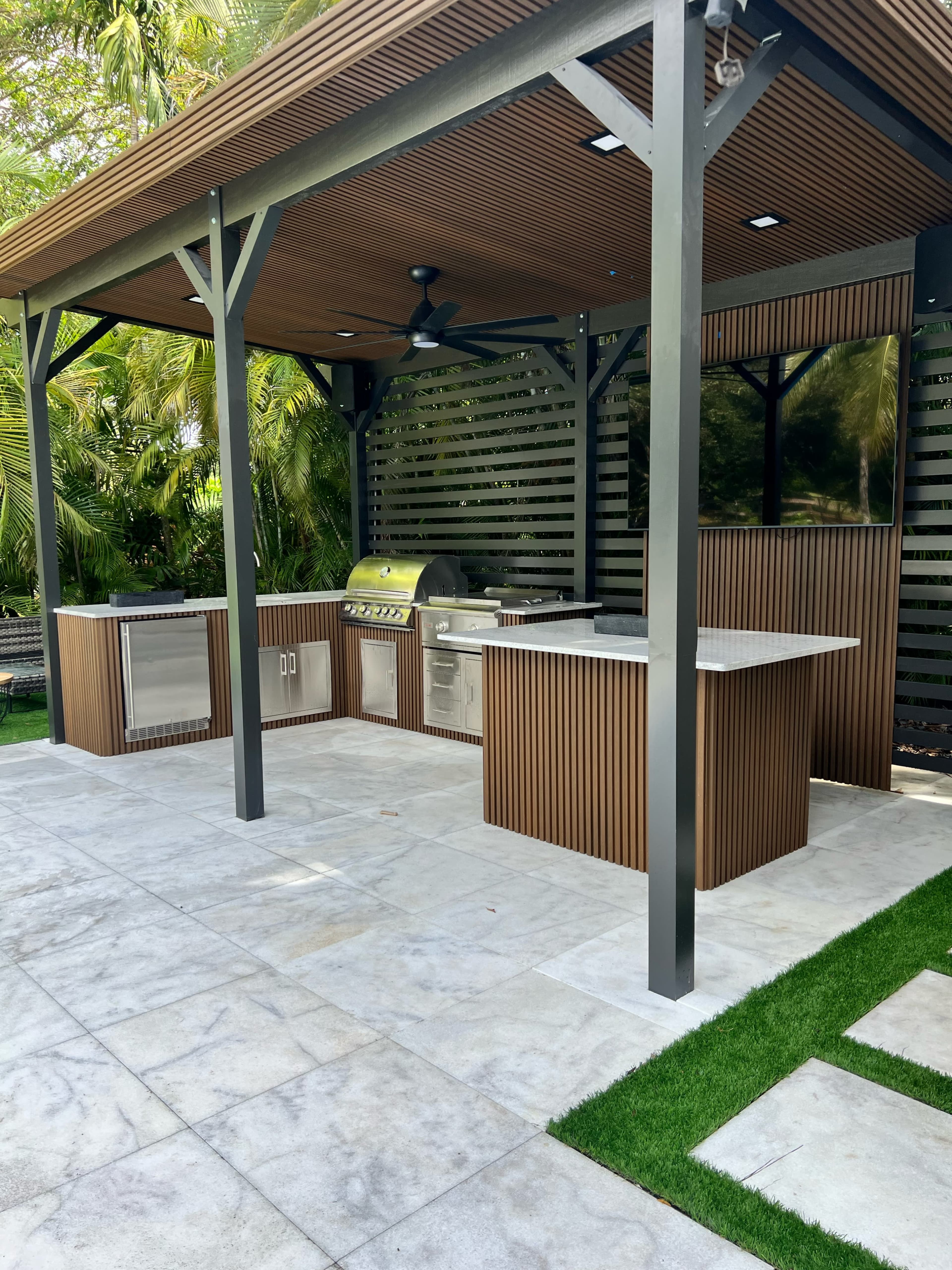 The image shows an outdoor kitchen pavilion featuring a grill, countertops, storage cabinets, and a television, surrounded by greenery.