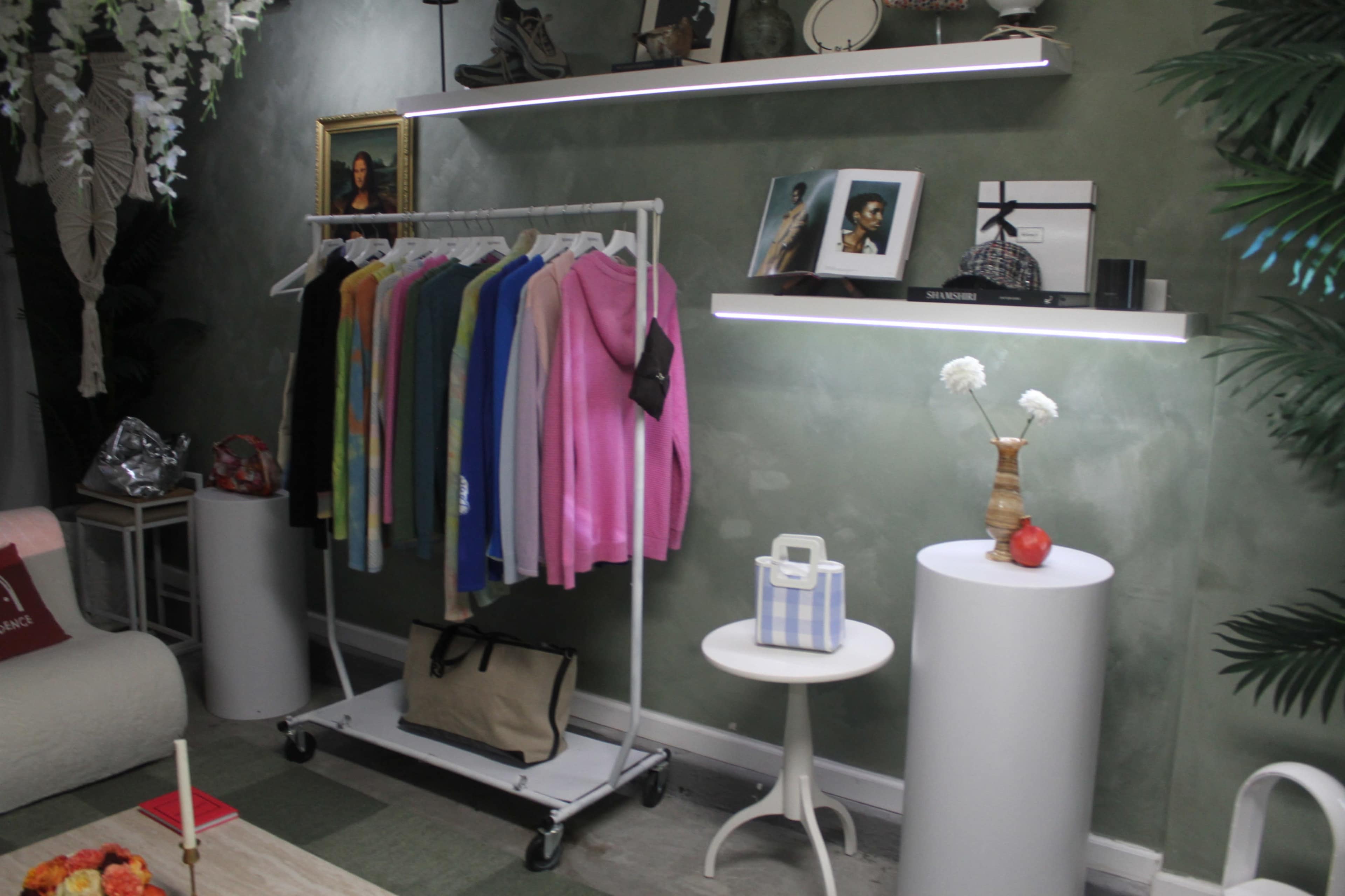 A clothing rack with various colorful garments is positioned next to a shelf displaying photos and decorative items in a stylish room with green walls.
