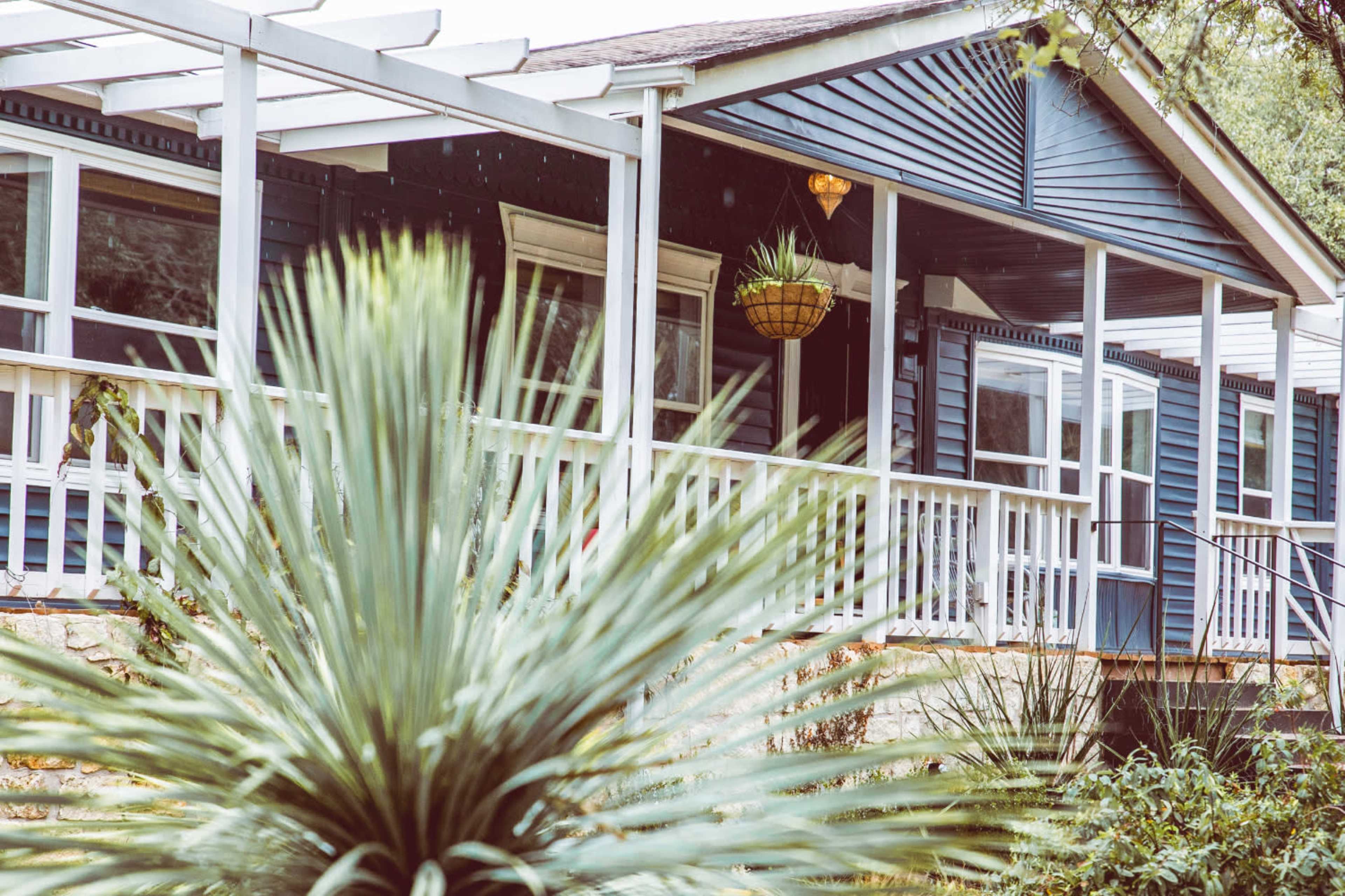 A blue and white house with a porch is surrounded by green plants under overcast skies.