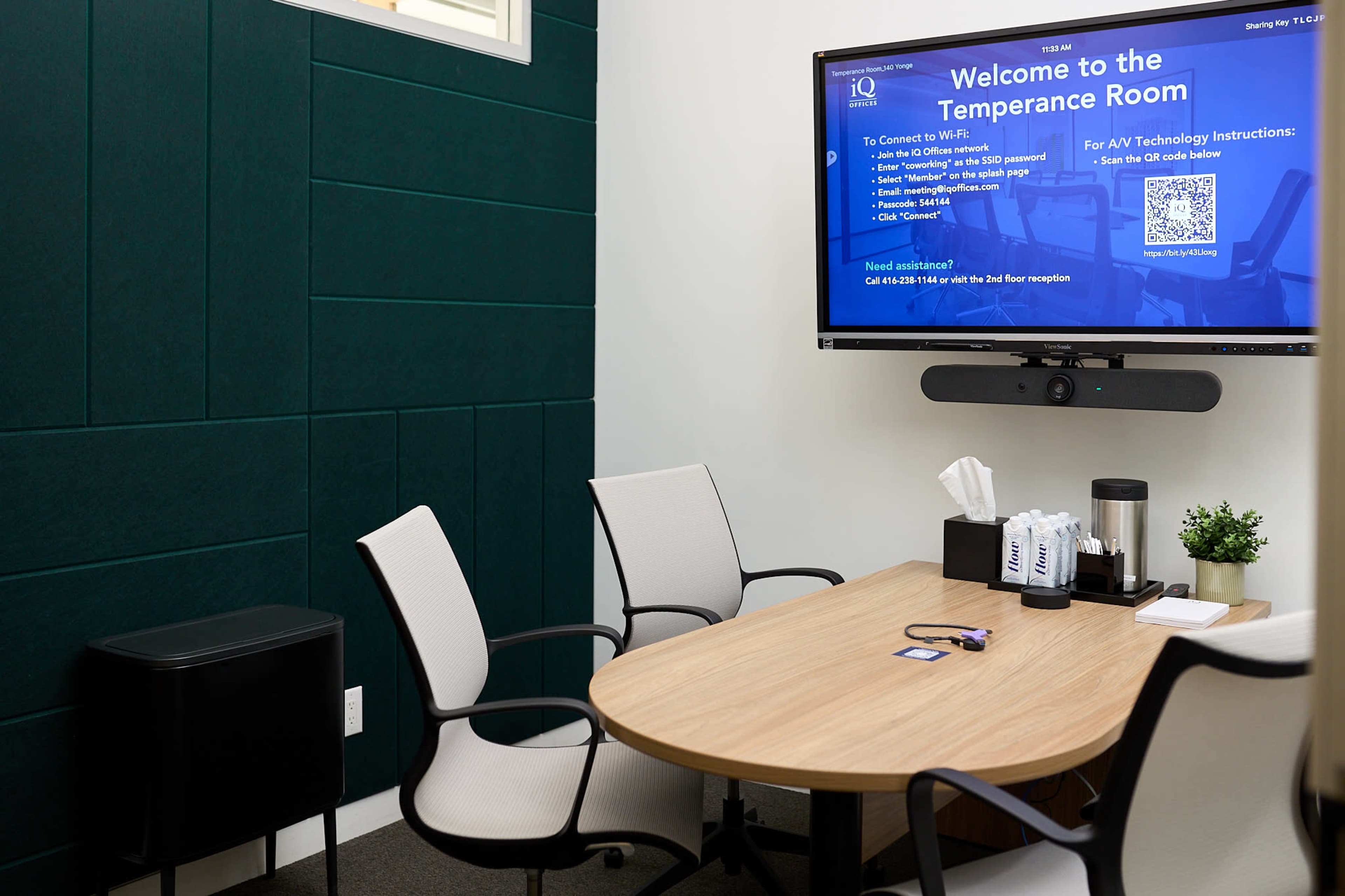 The image shows a small meeting room with a round wooden table, four chairs, a wall-mounted screen displaying instructions, and a green accent wall.