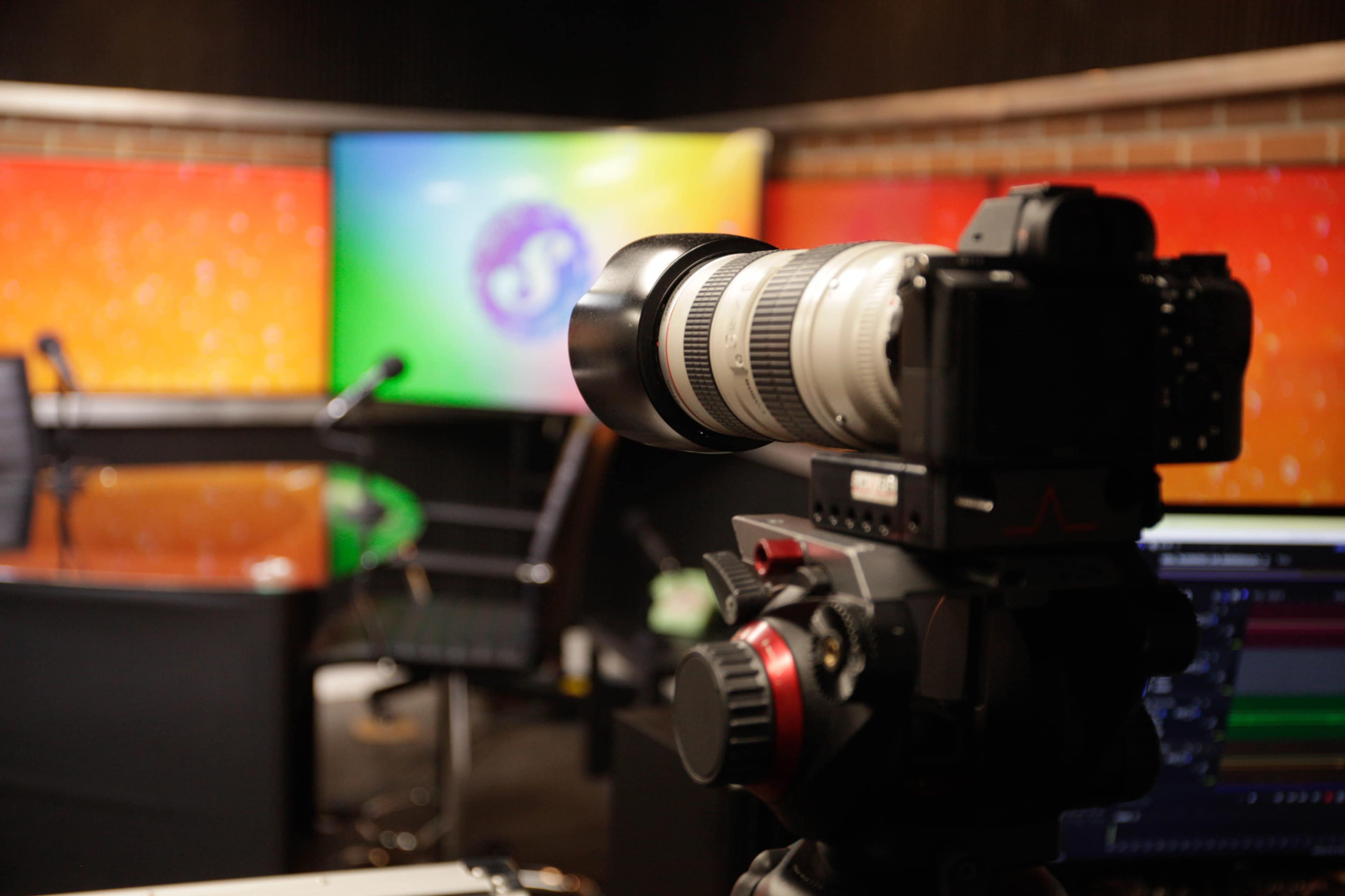 A camera is positioned on a tripod in a broadcasting studio with a colorful backdrop and a monitor displaying a logo.
