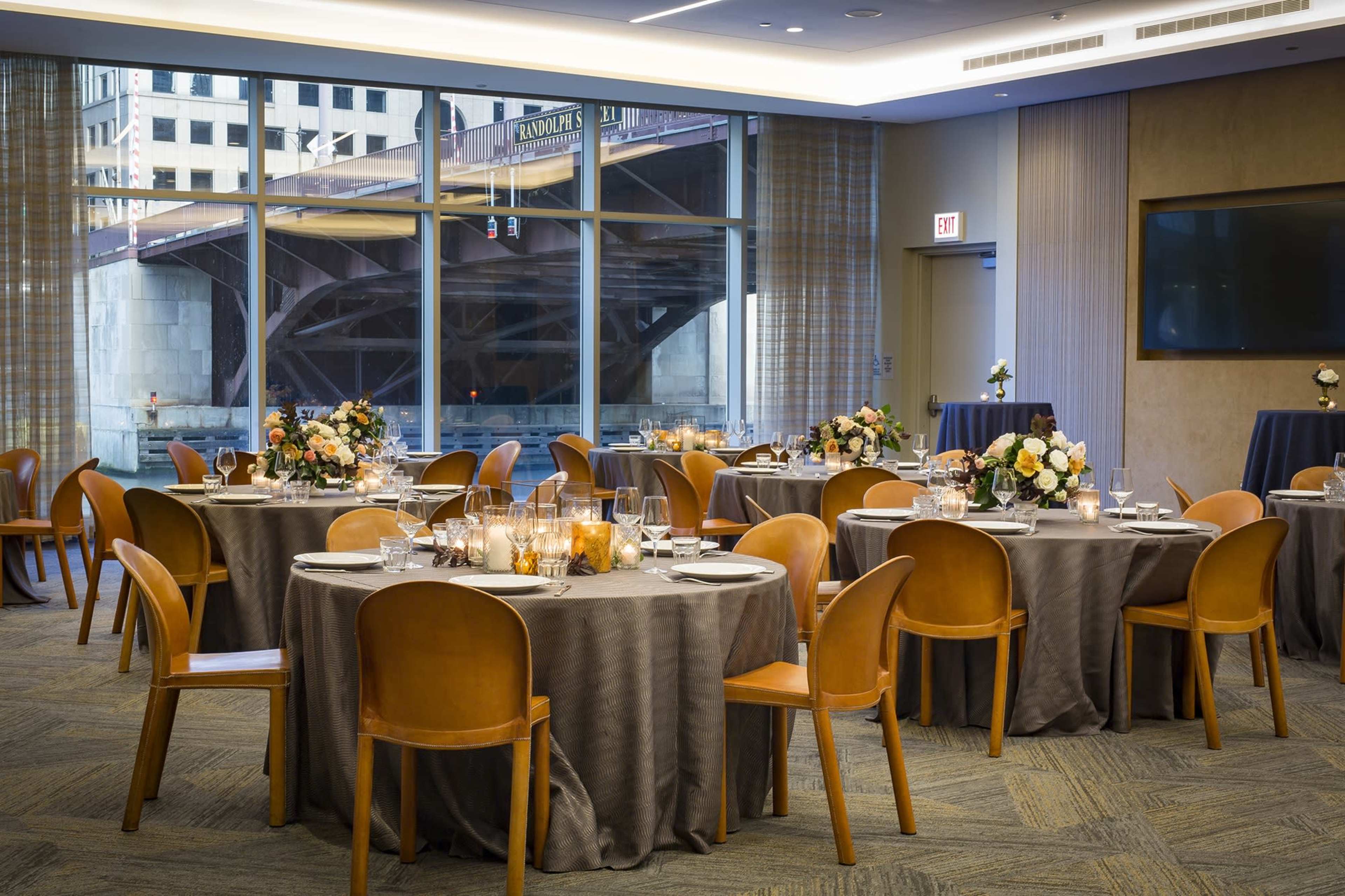 A dining room with several round tables set for a meal, featuring tablecloths, flower arrangements, and a view of a bridge through large windows.