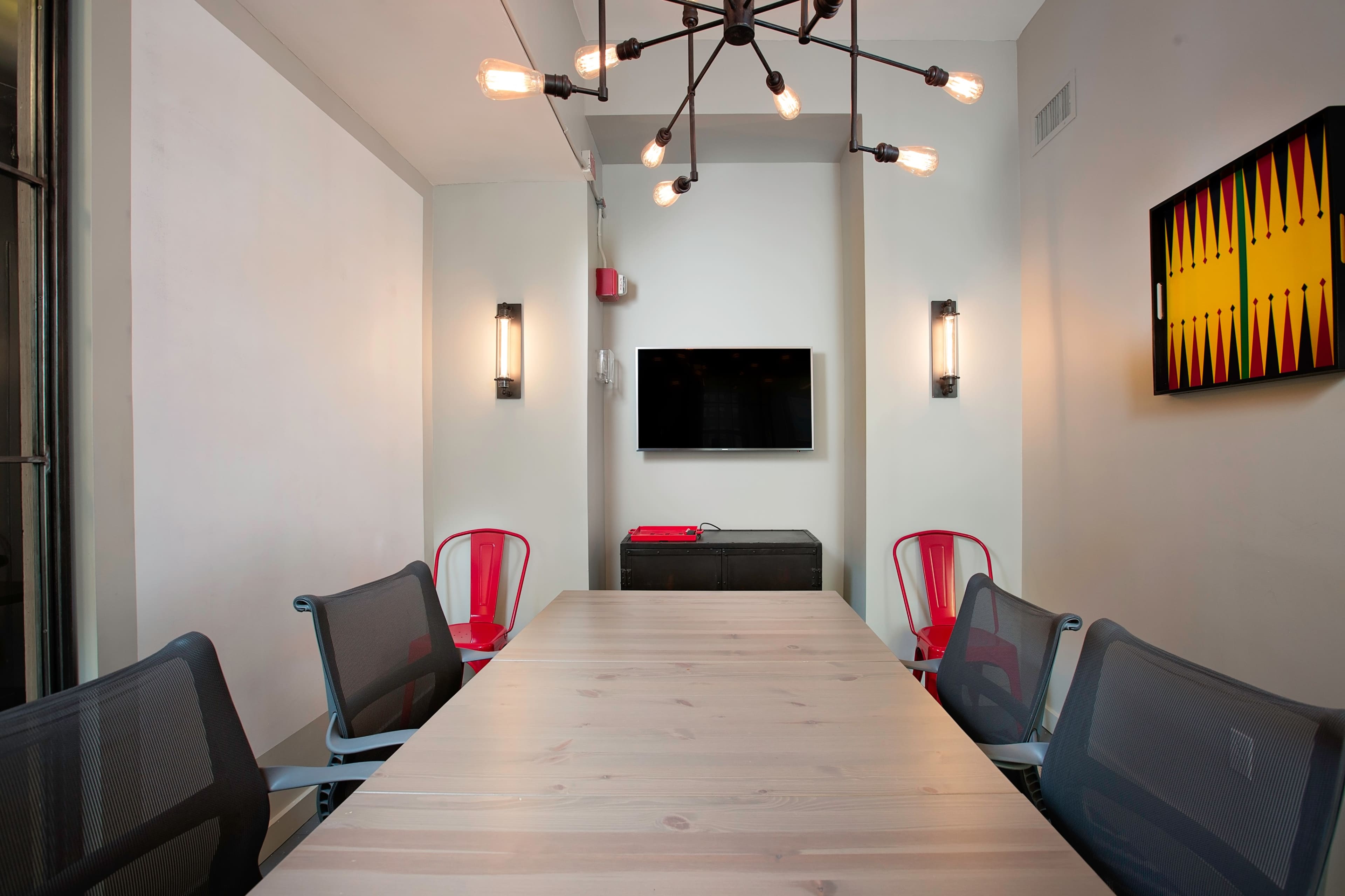 The image showcases a modern conference room with a long wooden table, four mesh chairs, a wall-mounted television, and a decorative wall piece.