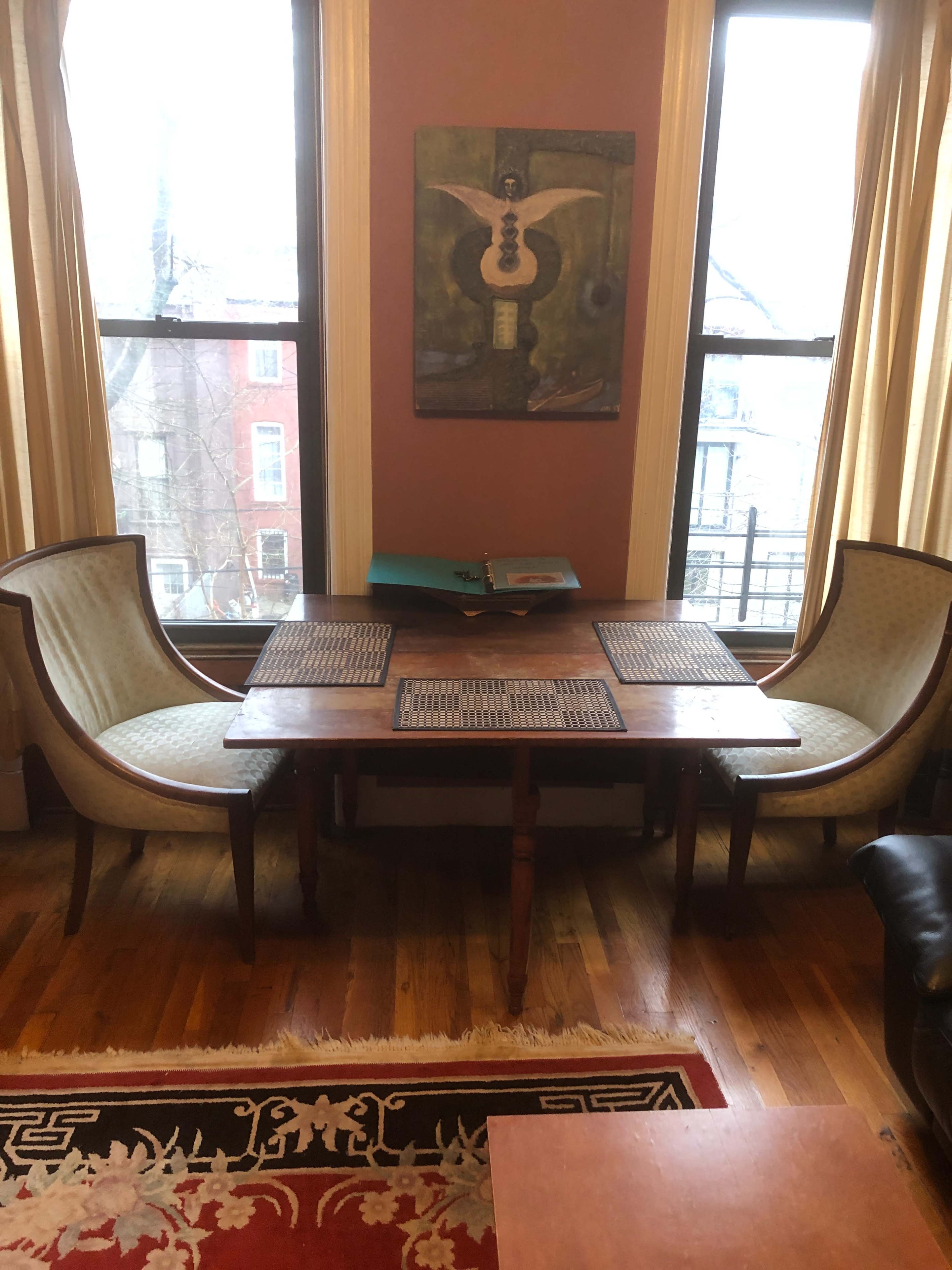 A wooden table is set between two upholstered chairs under large windows, with a painting hanging on the wall above.