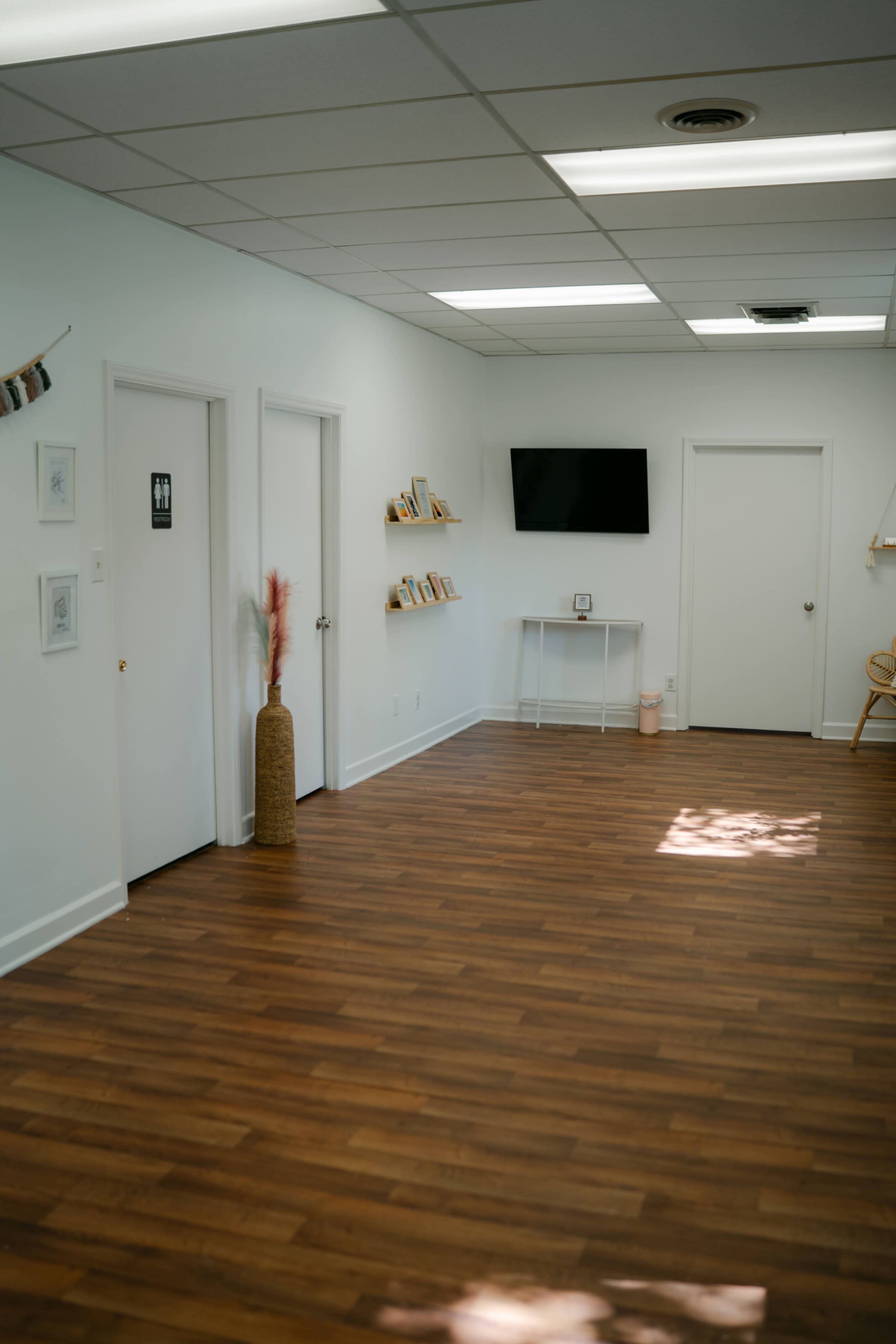 The image shows a spacious, minimally decorated room with wooden flooring, featuring multiple doors, a television mounted on the wall, and shelves with framed photos.