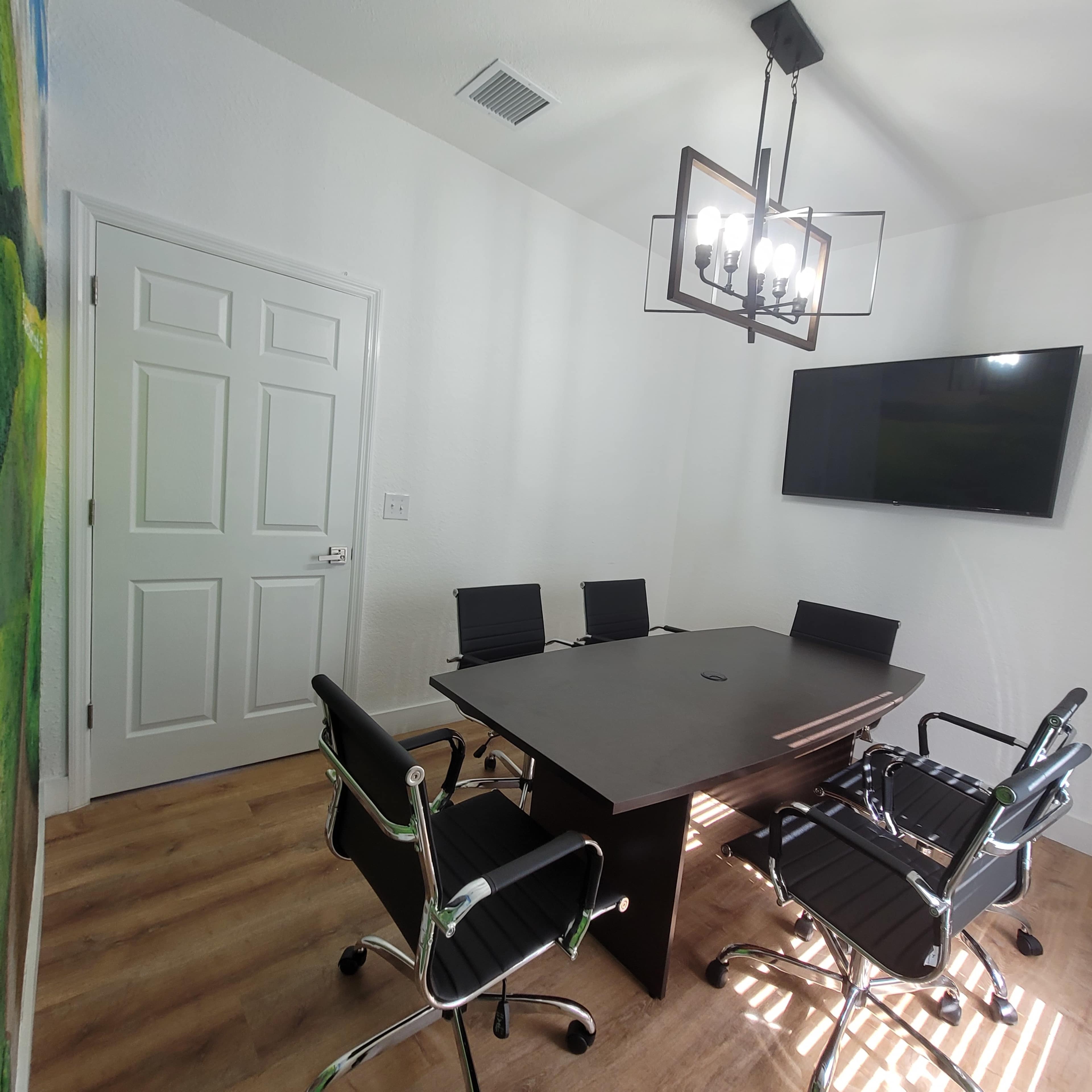 A modern conference room features a rectangular table surrounded by black chairs, a television mounted on the wall, and a chandelier overhead.