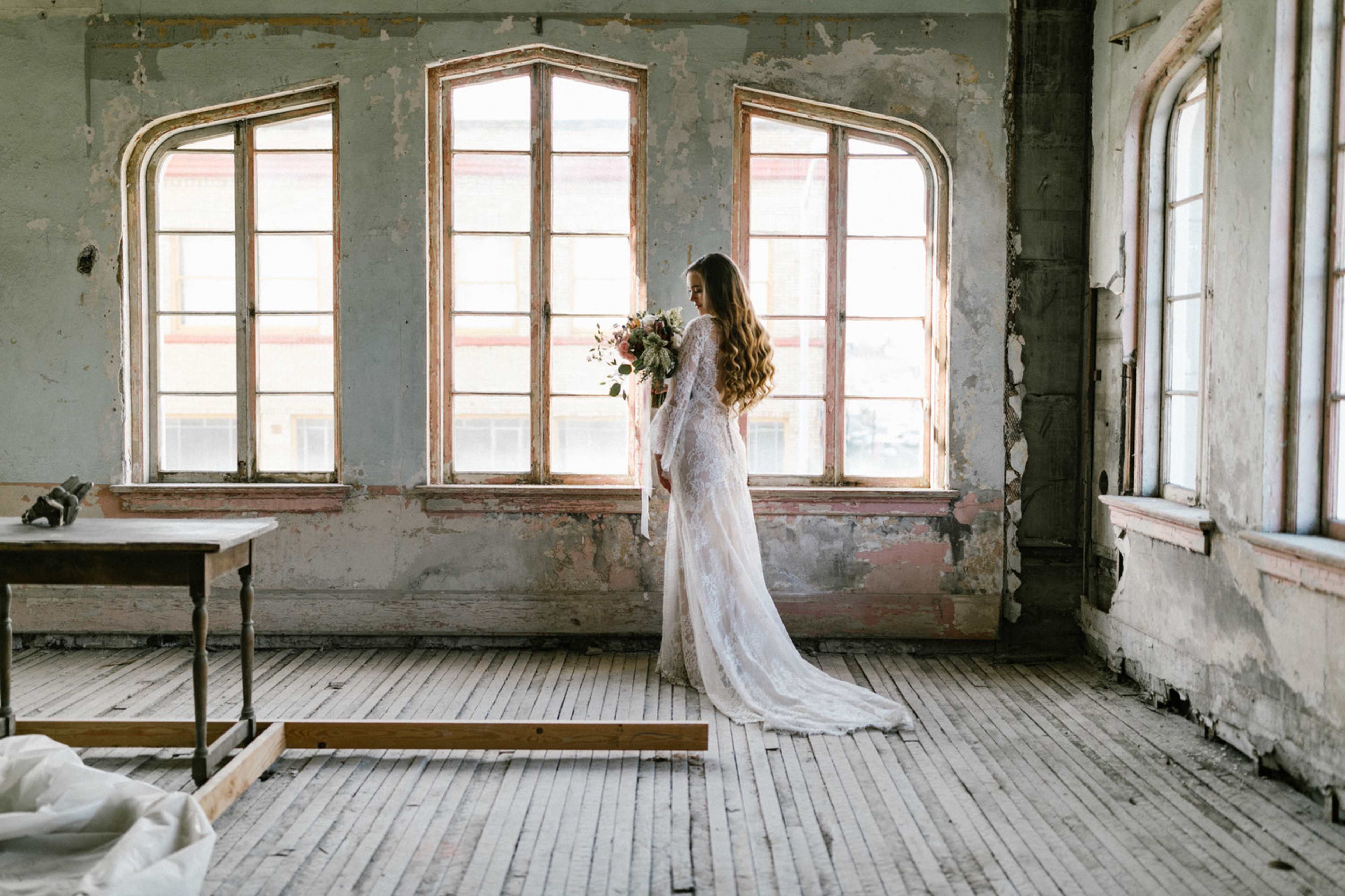 A woman in a white wedding dress stands with a bouquet of flowers in front of large, arched windows in a weathered, abandoned room.