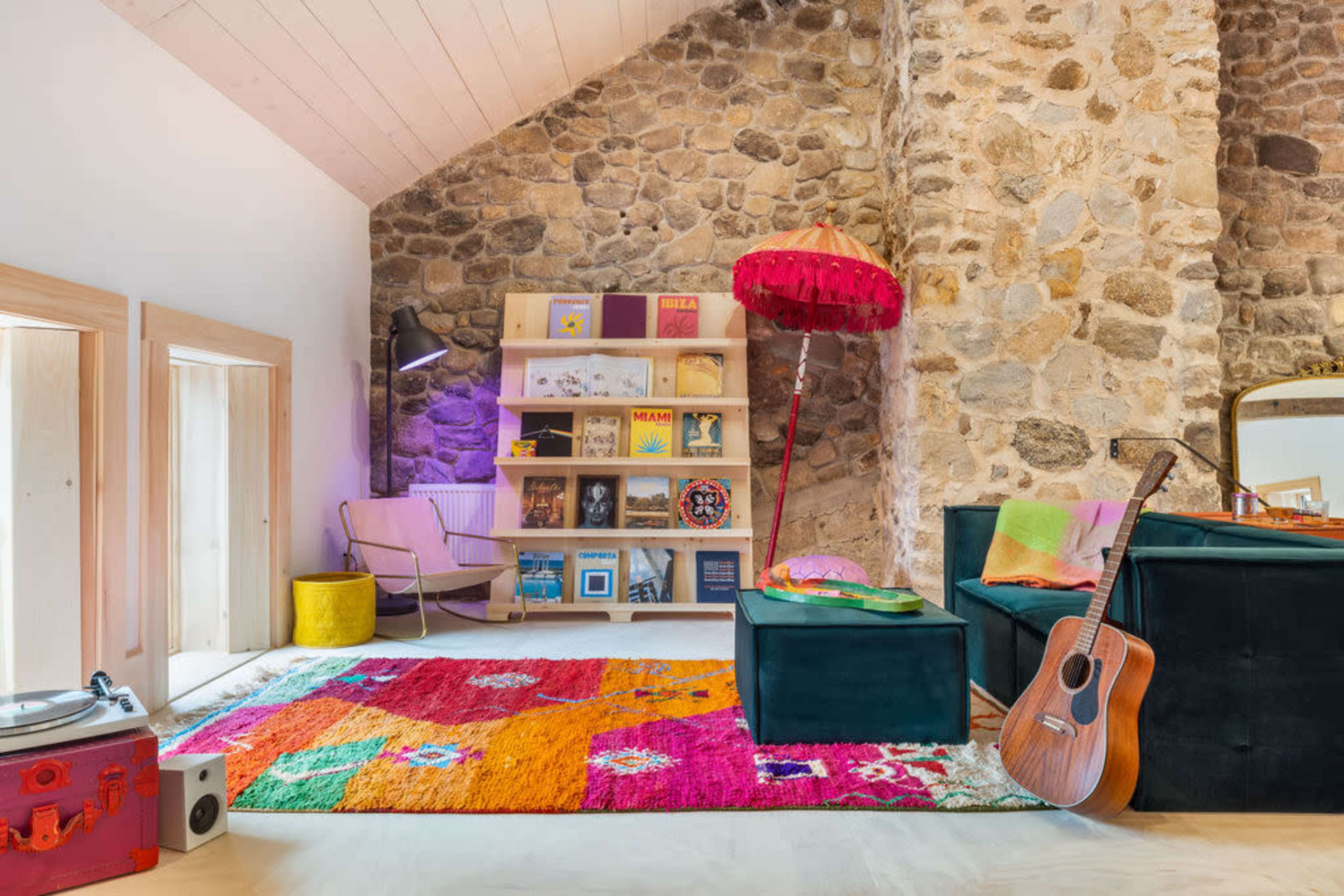 A living room with a stone wall, a bookshelf filled with colorful items, a patterned rug, a guitar, and a floor lamp.