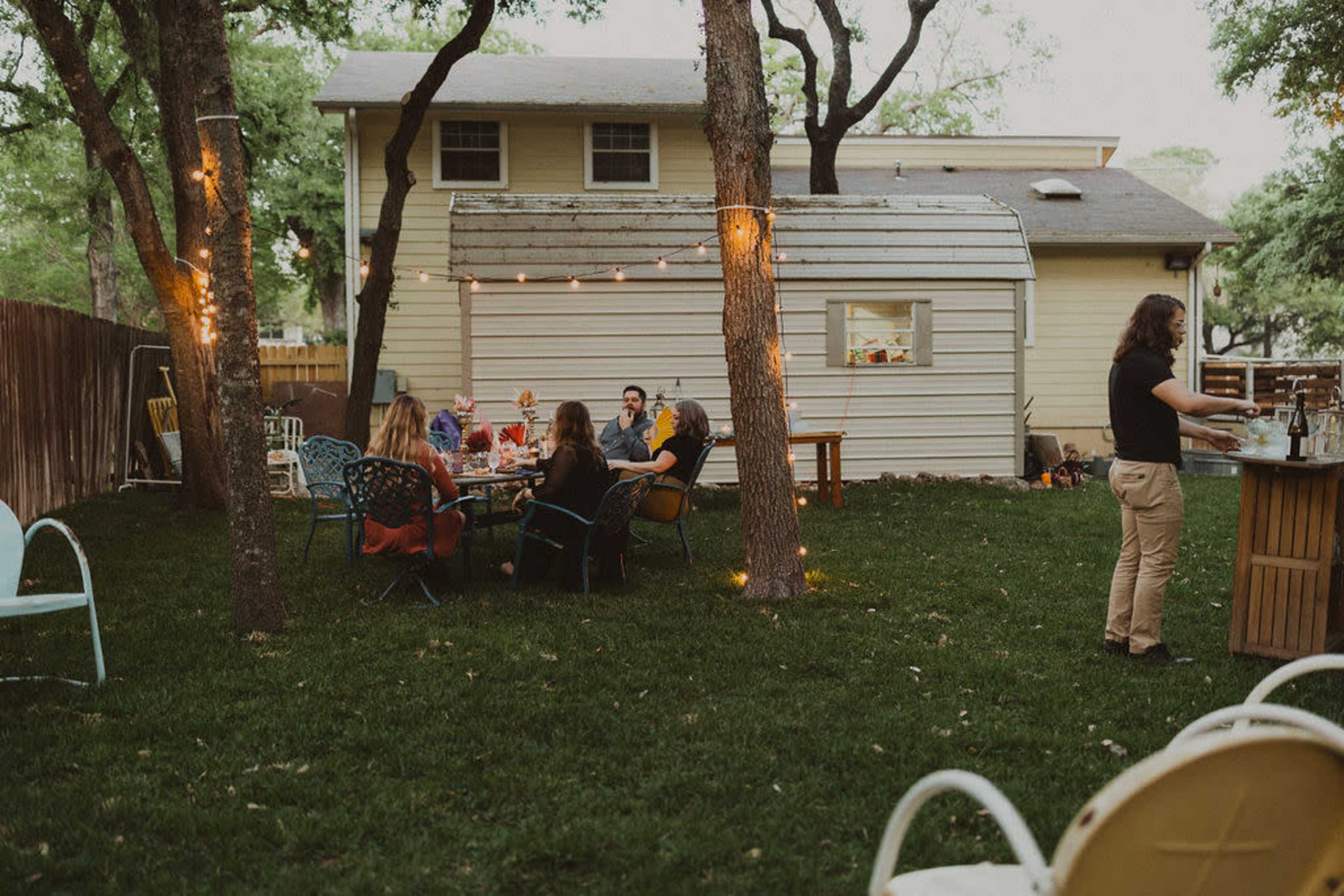 A group of people sits at a table in a backyard garden while another person tends to a bar nearby, illuminated by string lights and surrounded by trees.