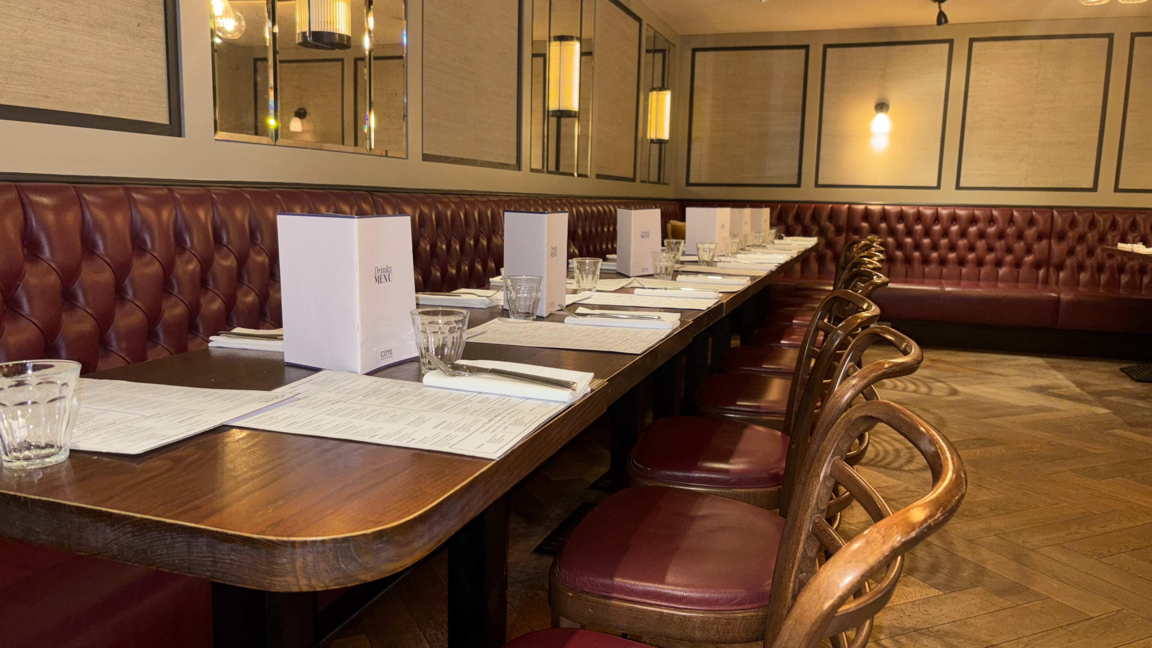 A long, empty dining table is set with menus and glasses in a restaurant with leather booth seating and pendant lights.