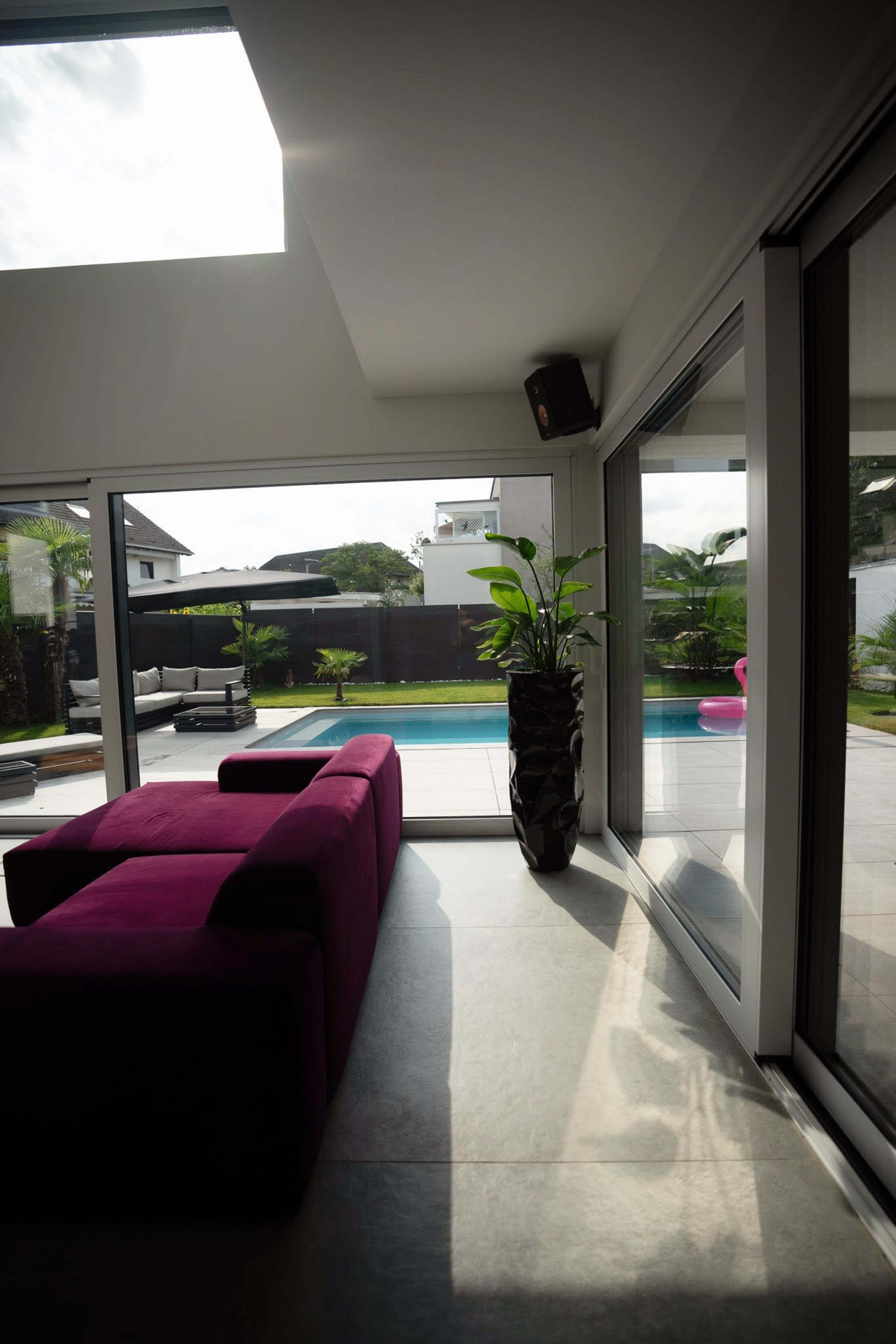 The image shows a modern living space with a purple couch, large glass doors leading to a pool area, and a tall potted plant.