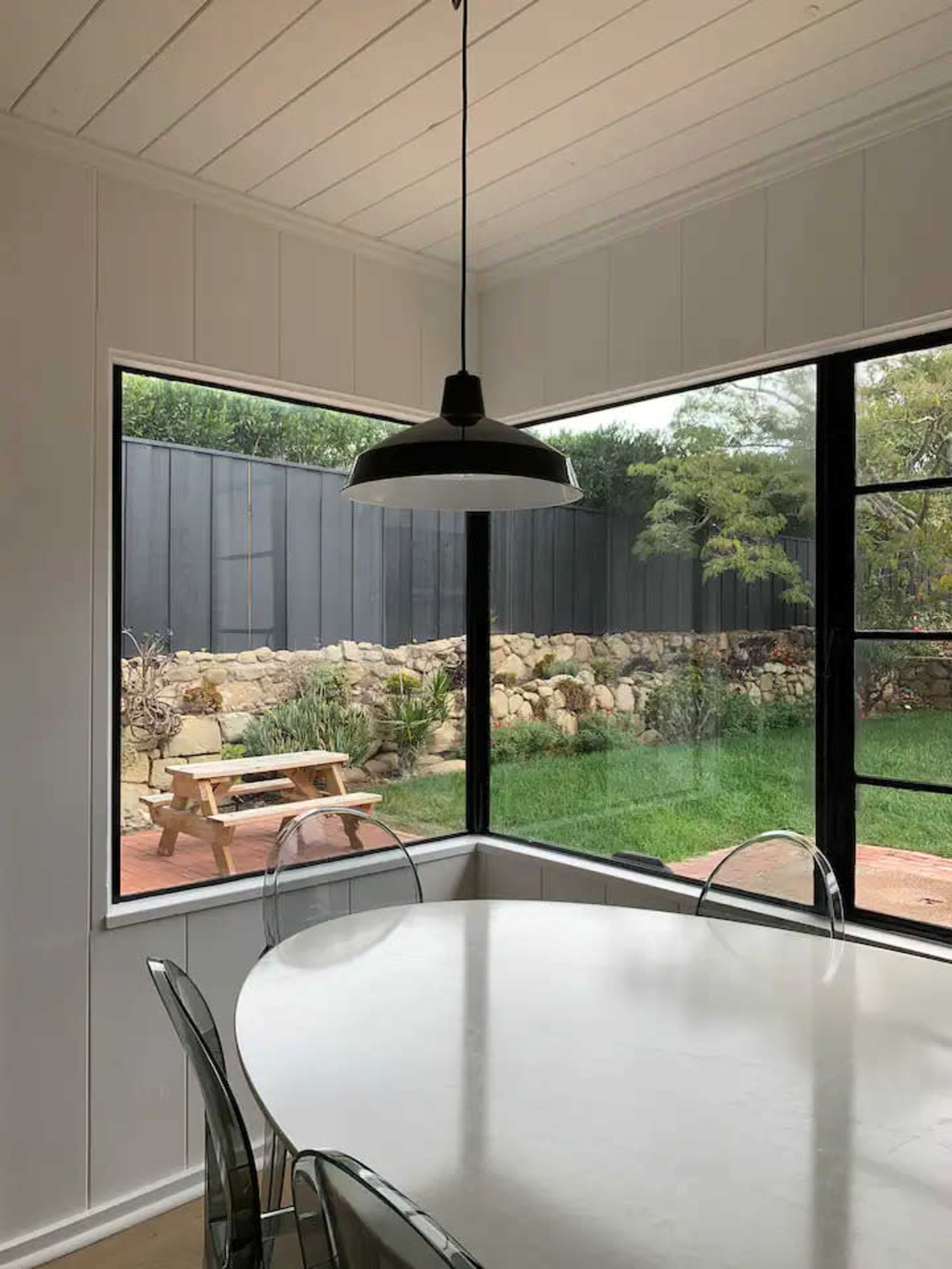 A dining area features a round white table surrounded by transparent chairs, with large windows offering a view of a garden and stone wall outside.