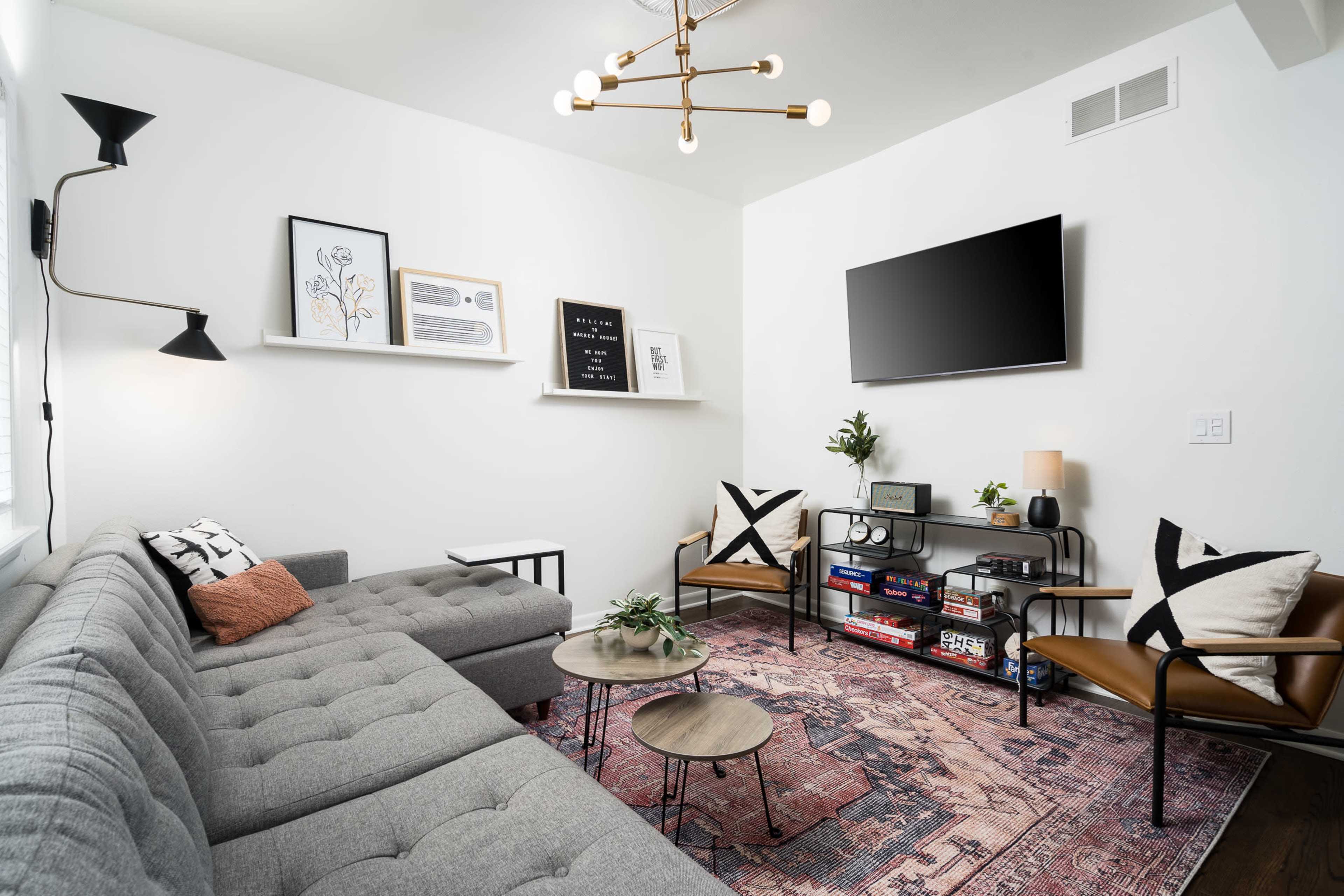 A cozy living room features a gray sectional sofa, mid-century chairs, a patterned rug, and various decorative elements on the walls and shelves.