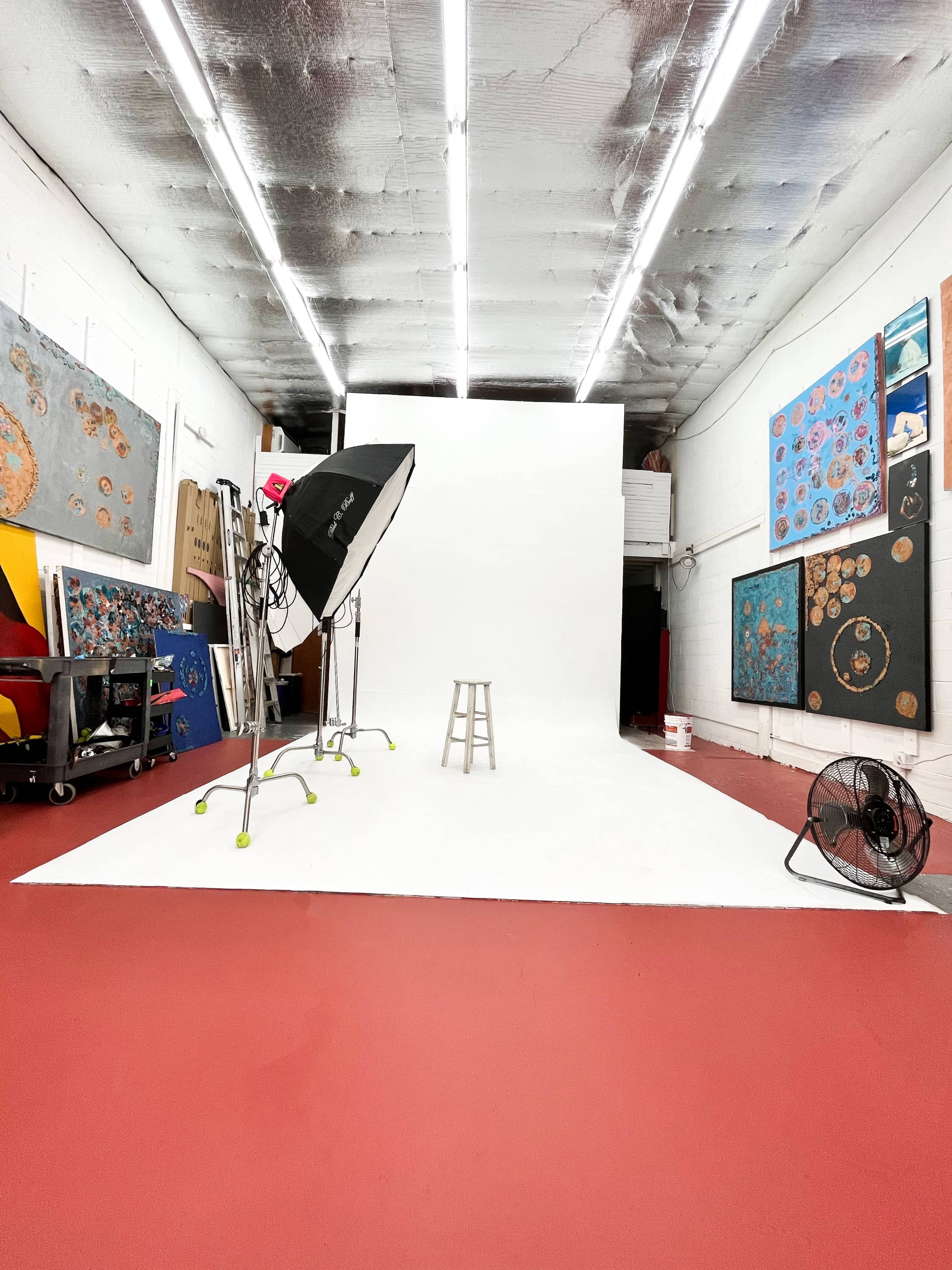 A photo studio with a white backdrop, a stool positioned in front, lighting equipment, and artwork displayed on the walls.
