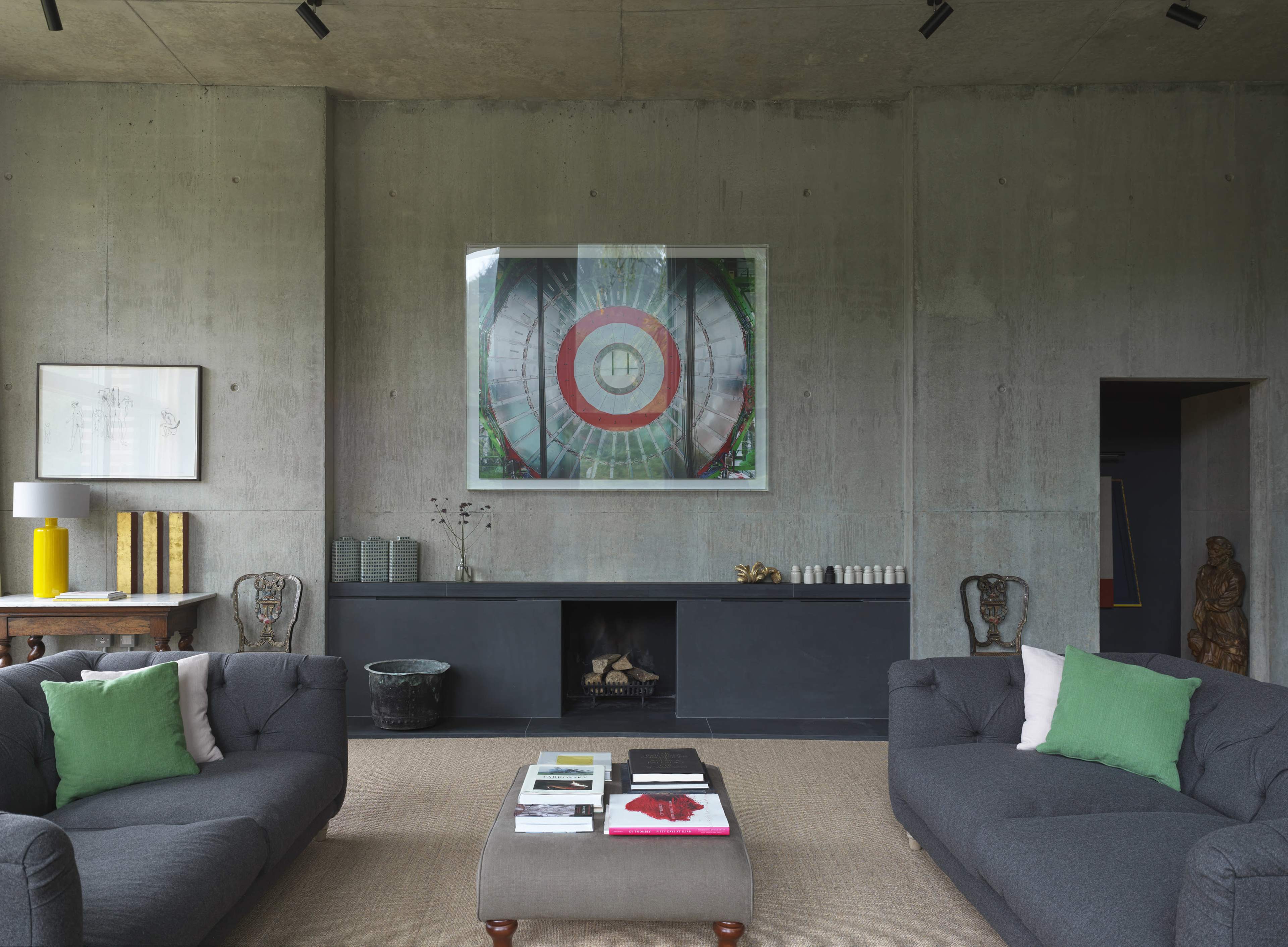 A modern living room features two gray sofas with green cushions, a coffee table with various books, and a large artwork on the concrete wall above a minimalist black console.