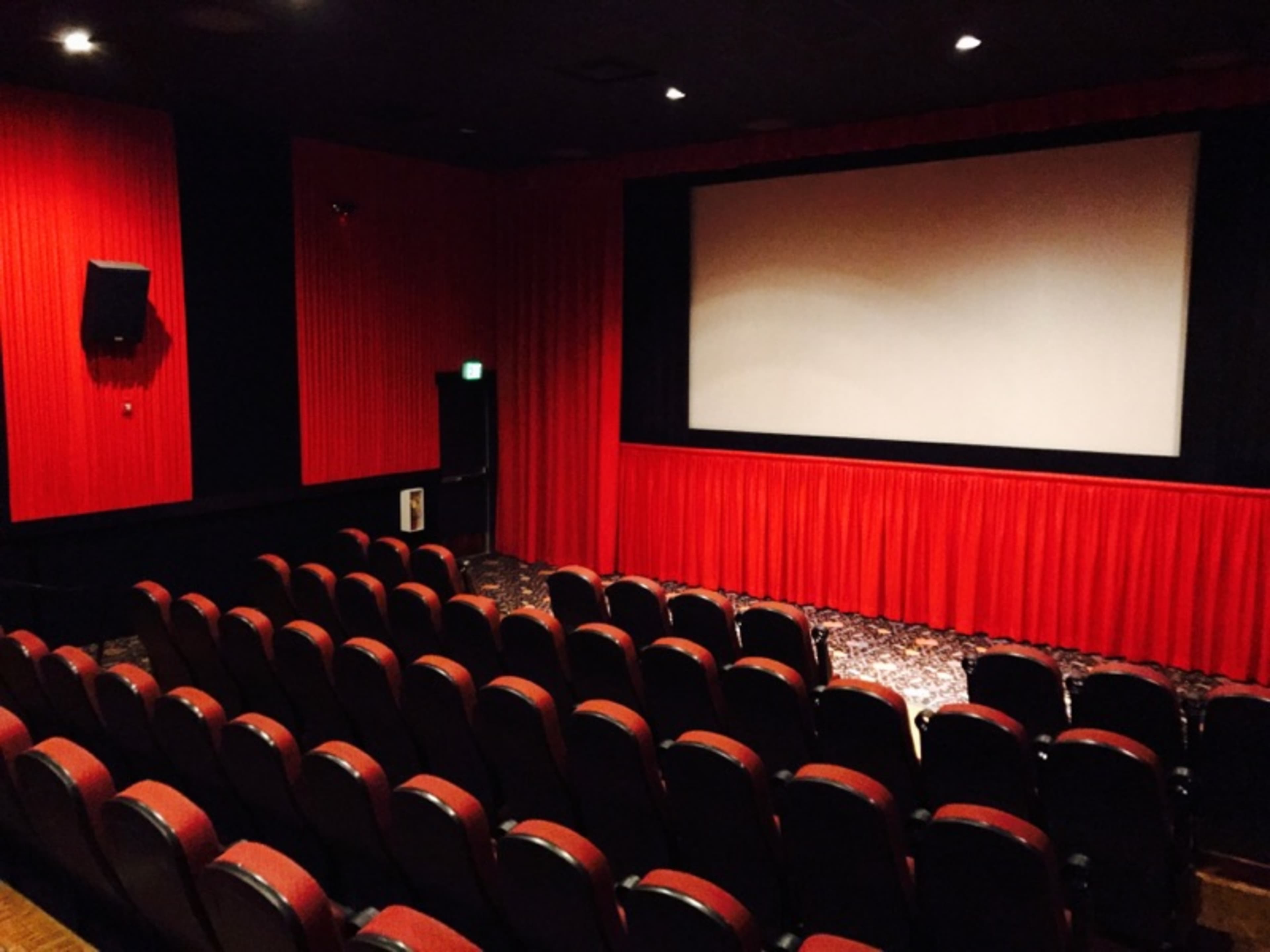 An empty movie theater with red curtains and rows of black seats facing a blank screen.