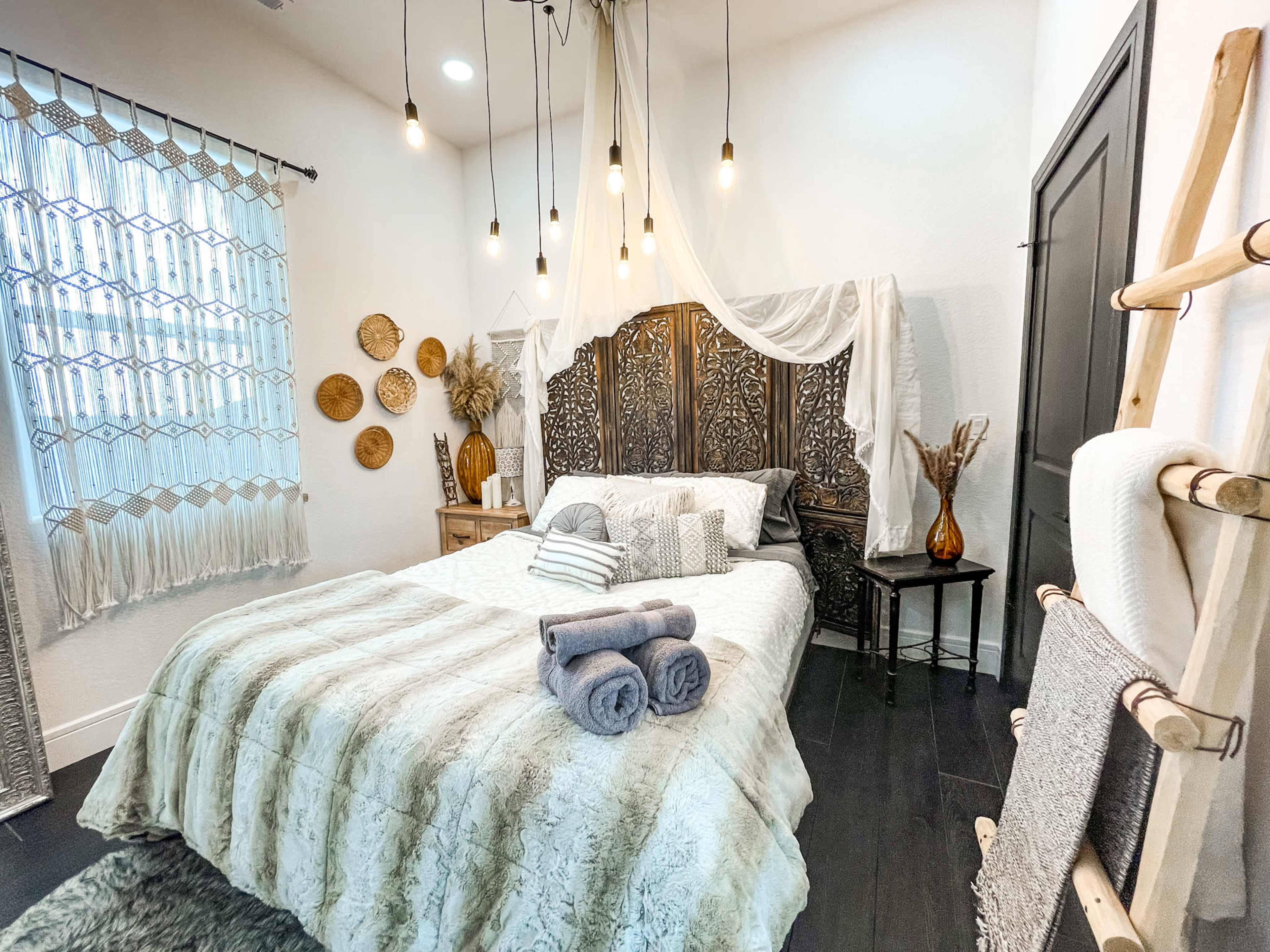 The scene features a styled bedroom with a large bed, a decorative headboard, patterned bedding, and ambient lighting from hanging bulbs.