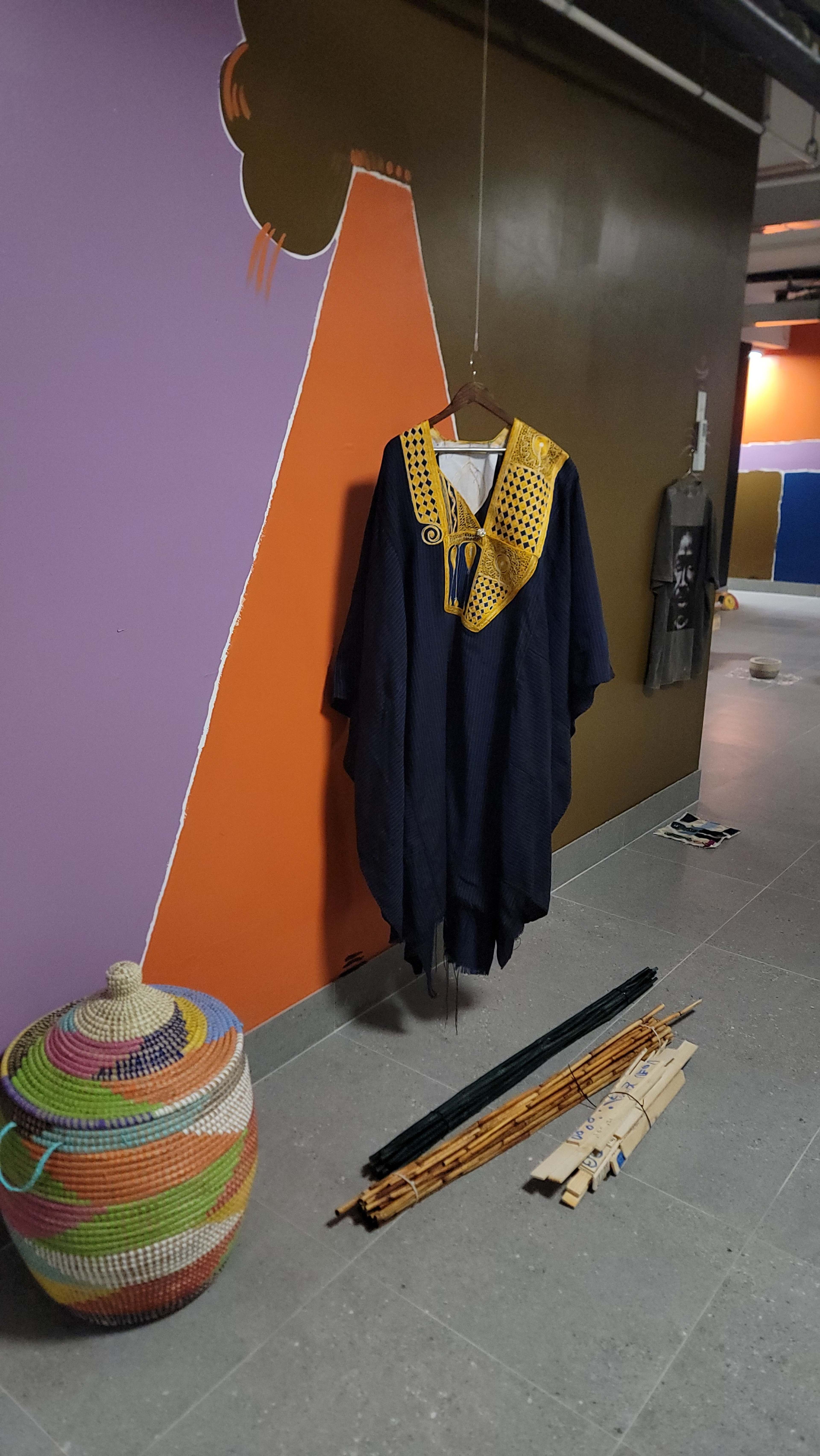 The image shows a traditional garment with a distinct yellow neck design hanging on a wall, accompanied by a colorful woven basket and some wooden items on the floor.