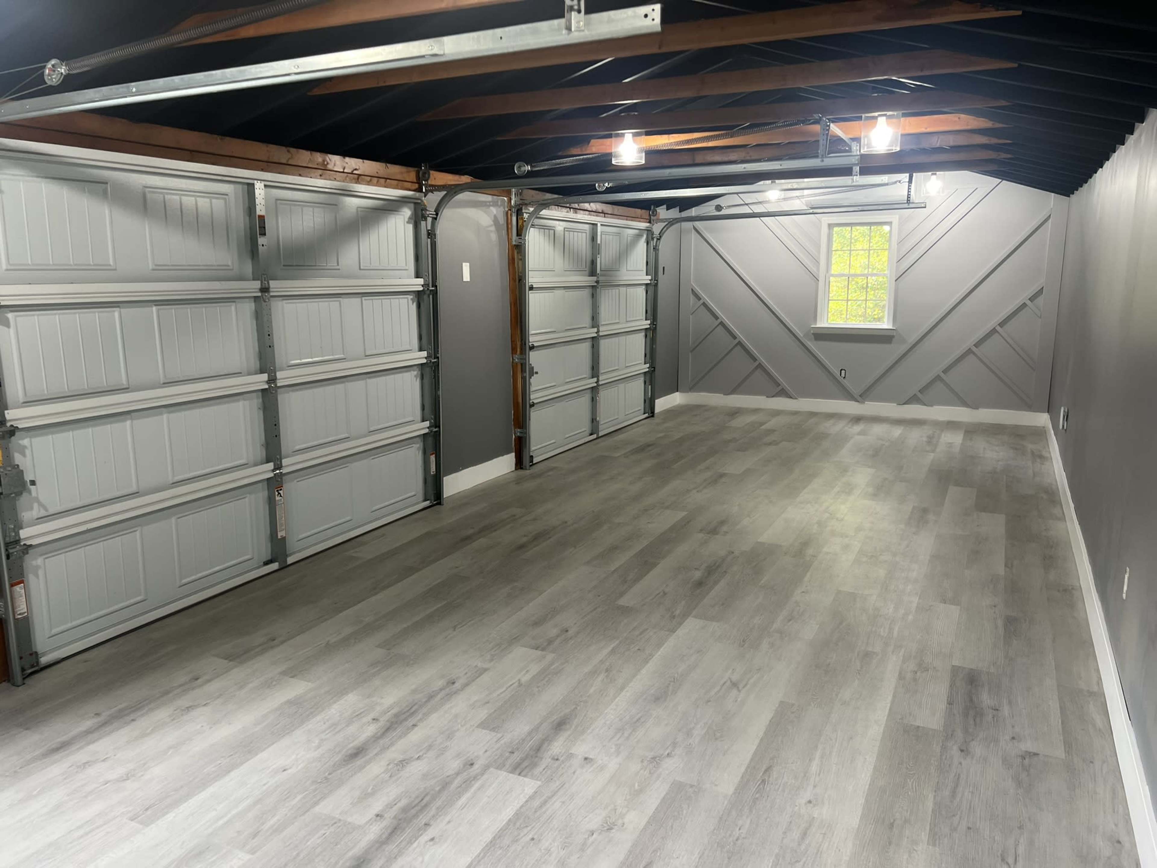 An empty garage with two closed garage doors and a window, featuring gray walls and a light-colored wood floor.