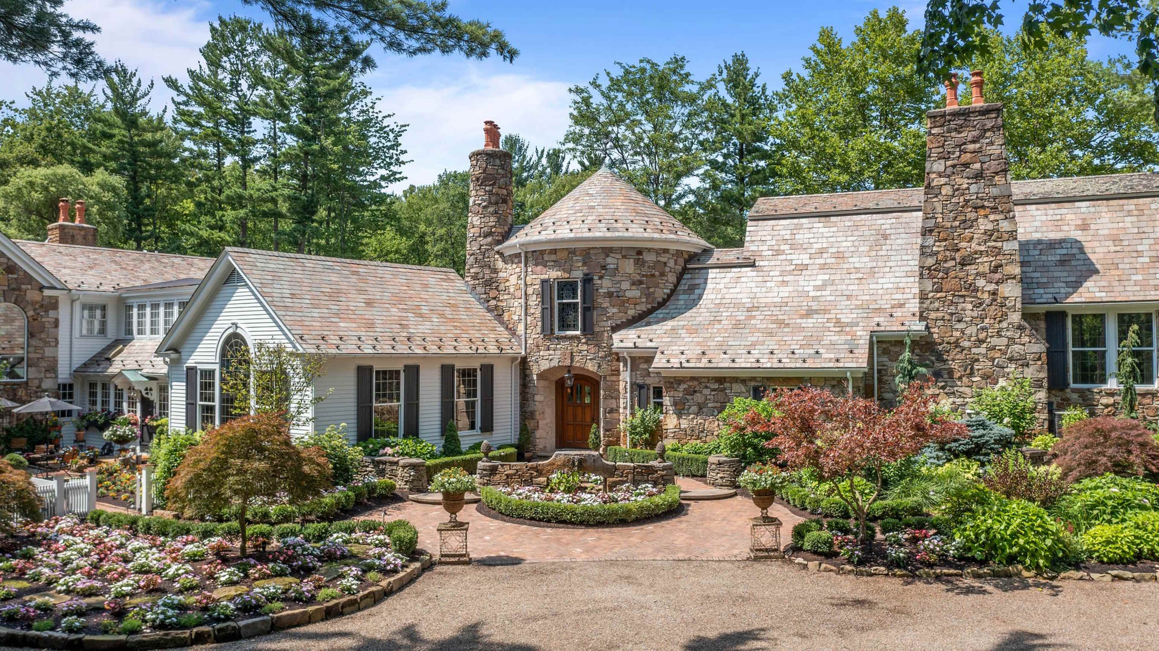 The image features a stone and shingle house with a circular tower and landscaped garden filled with colorful flowers and shrubs.