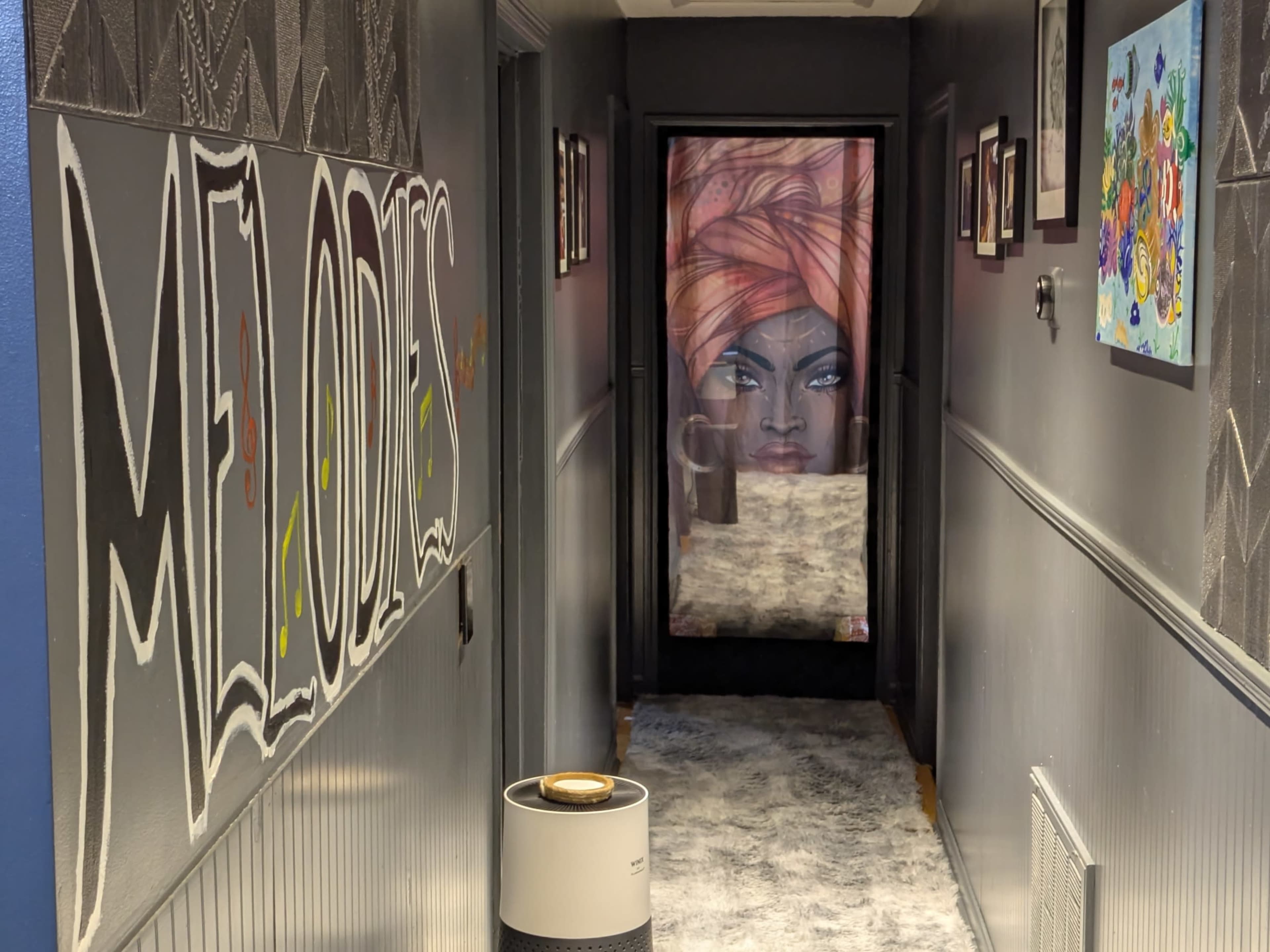 The image shows a narrow hallway with colorful murals on the walls and a large mirror at the far end reflecting a portrait.