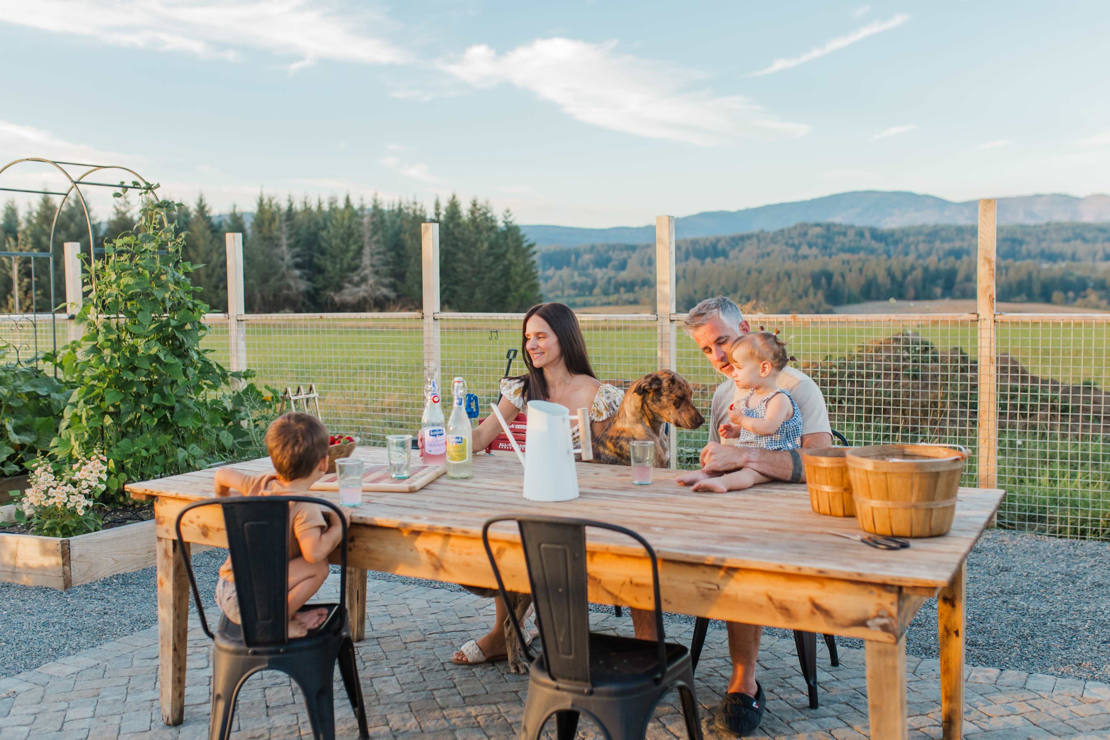 A family is sitting around a wooden table outdoors, enjoying drinks and snacks with a dog, set against a backdrop of green fields and mountains.