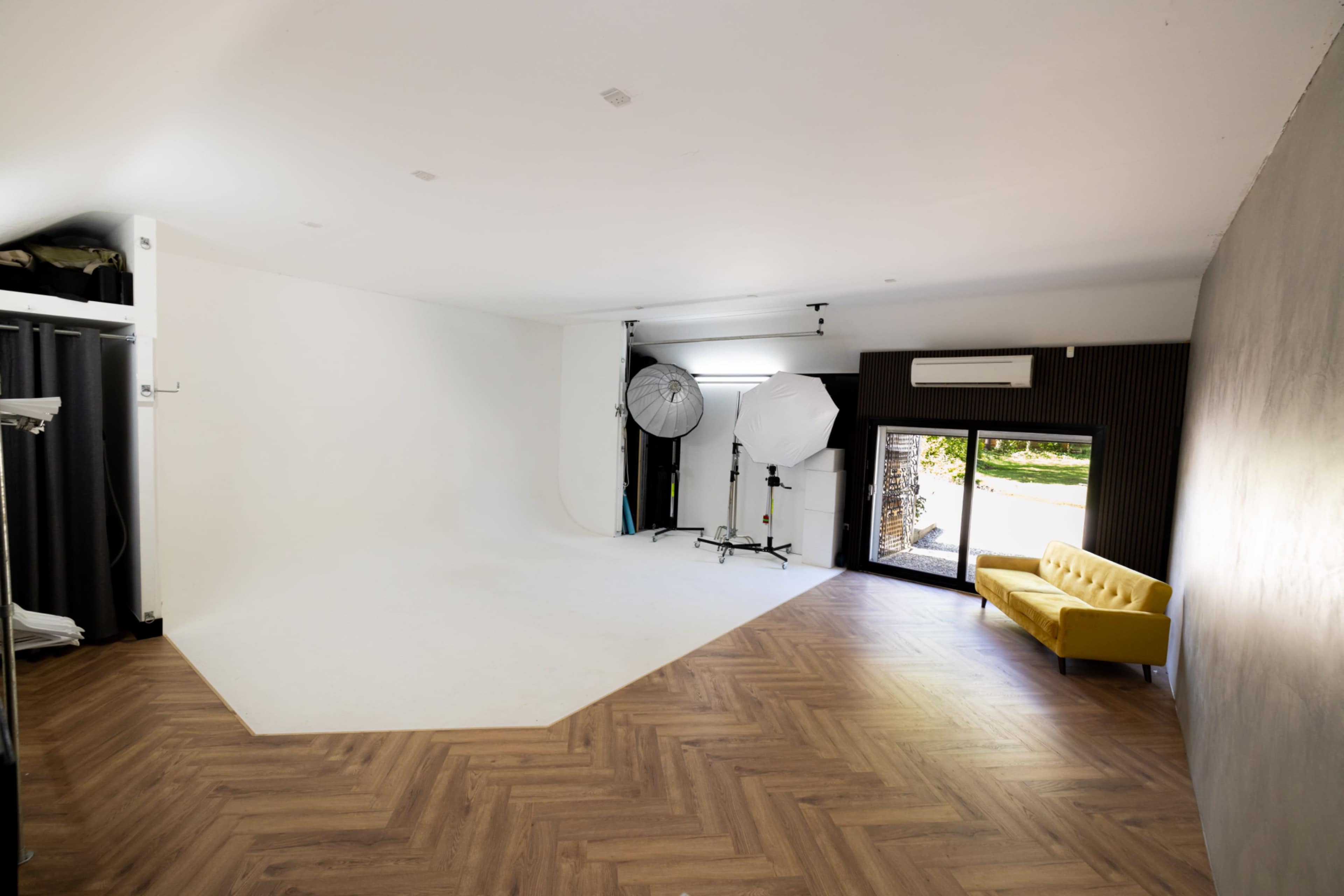 A photography studio featuring a large white backdrop, lighting equipment on tripods, and a yellow sofa against a wooden floor.