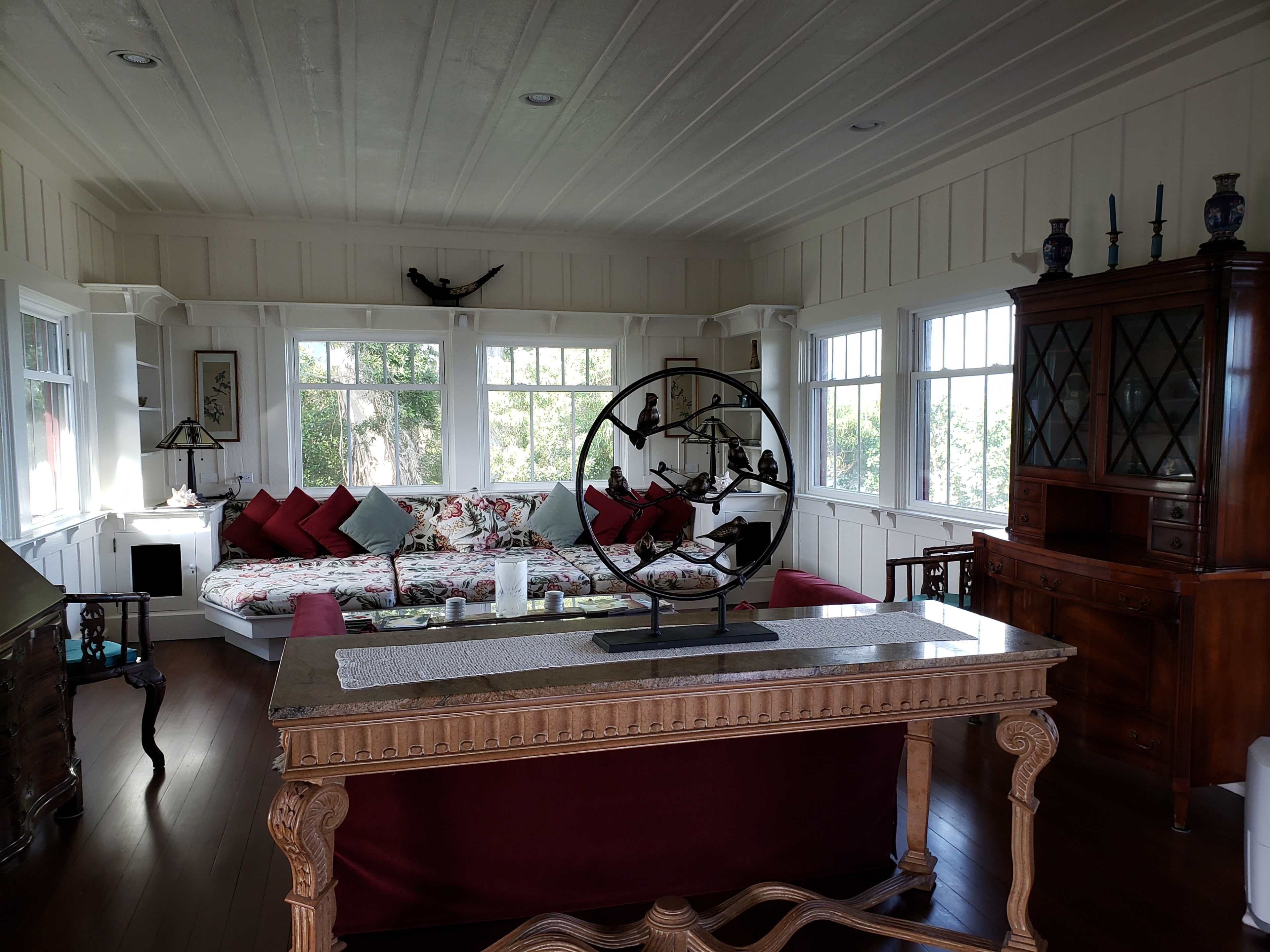The image shows a spacious living room with large windows, a floral-patterned sofa, a wooden dining table, and a decorative display cabinet.