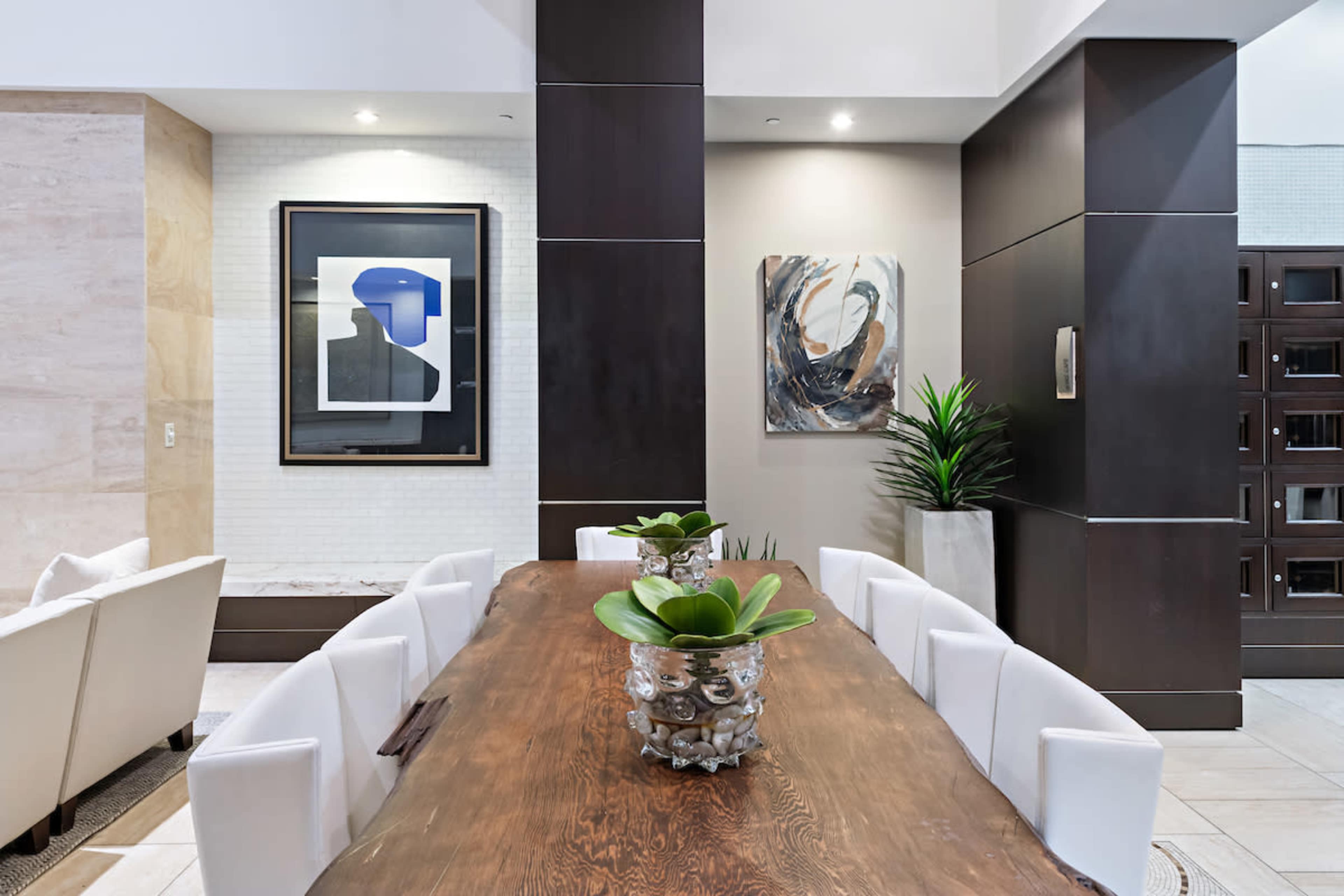 A modern dining area features a large wooden table surrounded by white chairs, with decorative plants and artwork on the walls.